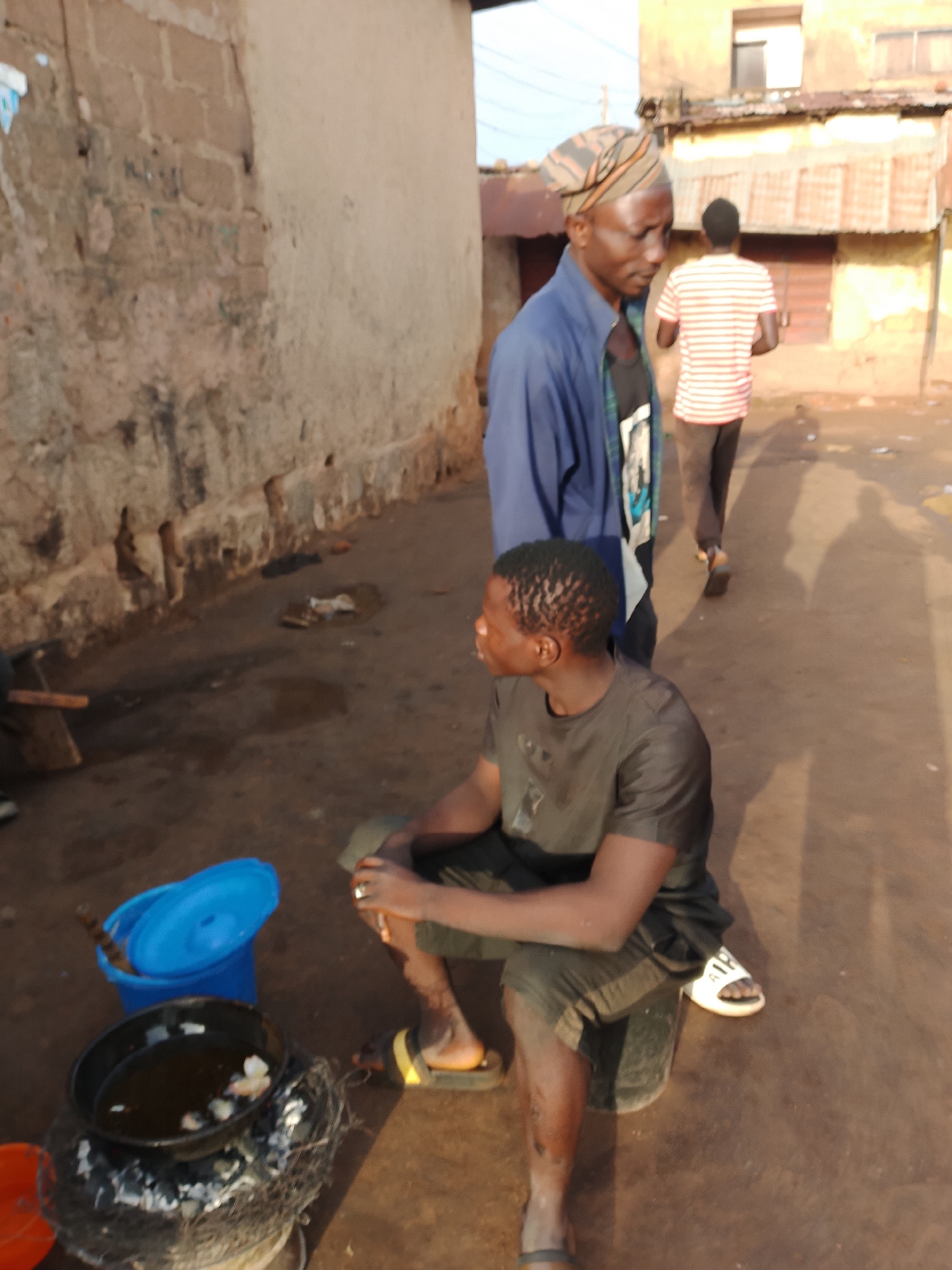 In Jos, Nigeria, two men are present in an outdoor urban environment. The man in the foreground, seated, wears a dark t-shirt and dark shorts, with white slides on his feet. He is positioned next to a cooking setup that includes a pot of oil over burning coals, a blue bucket with a lid, and an orange bucket. The second man stands behind him, wearing a blue jacket over a black t-shirt and a patterned headwrap. In the background, another man in a striped shirt walks away, and buildings with corrugated metal roofs are visible. The ground is dirt and scattered with debris. The overall scene depicts a street-side cooking activity.