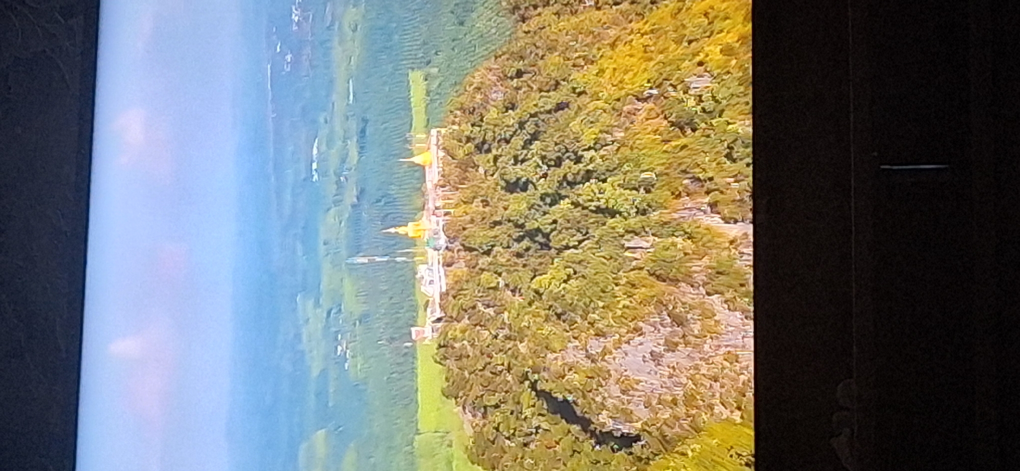 The image shows an aerial view of a monastery situated on a lush green mountain. The monastery complex features multiple golden stupas and white buildings. The surrounding landscape is a verdant expanse of trees and fields, with hints of structures and possibly roads in the distance. The sky is partly cloudy and appears to be a hazy blue. There is a dark border at the top and bottom of the image, suggesting it may be a photograph of a screen or display. The scene does not depict any people or specific actions. The location in the city of Bila Tserkva, Ukraine is not evident from the visual information provided in the image.