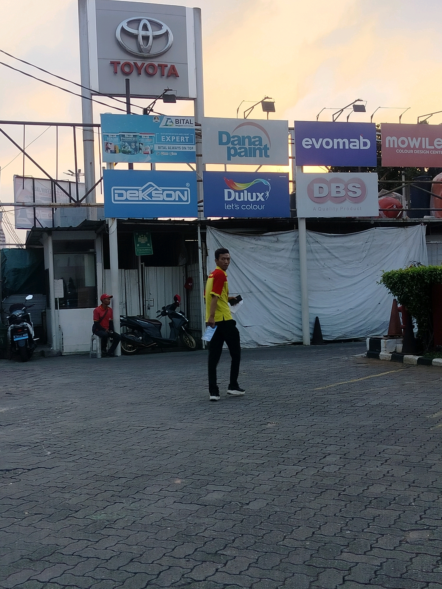 In East Jakarta, Indonesia, at a Toyota dealership, a street scene unfolds during what appears to be early morning or late afternoon, indicated by the warm, diffused light. A large, prominent Toyota sign, featuring the iconic logo and the word "TOYOTA" in red lettering, dominates the upper portion of the frame. Below the Toyota sign, a series of advertising billboards are displayed. These include advertisements for BITAL (waterproofing and paint), Dana Paint, evomab, Mowilex, Dekson, Dulux, and DBS.
In the foreground, a man wearing a red and yellow polo shirt and dark pants is walking away from the camera, holding a piece of paper. To the left, another man in a red shirt and red cap is seated on a white stool next to a small booth, appearing to be on duty. Two motorcycles are parked nearby. The ground is paved with interlocking dark grey bricks. A white tarp covers an area behind the billboards and booth. The overall impression is a commercial area during a quiet period of the day.