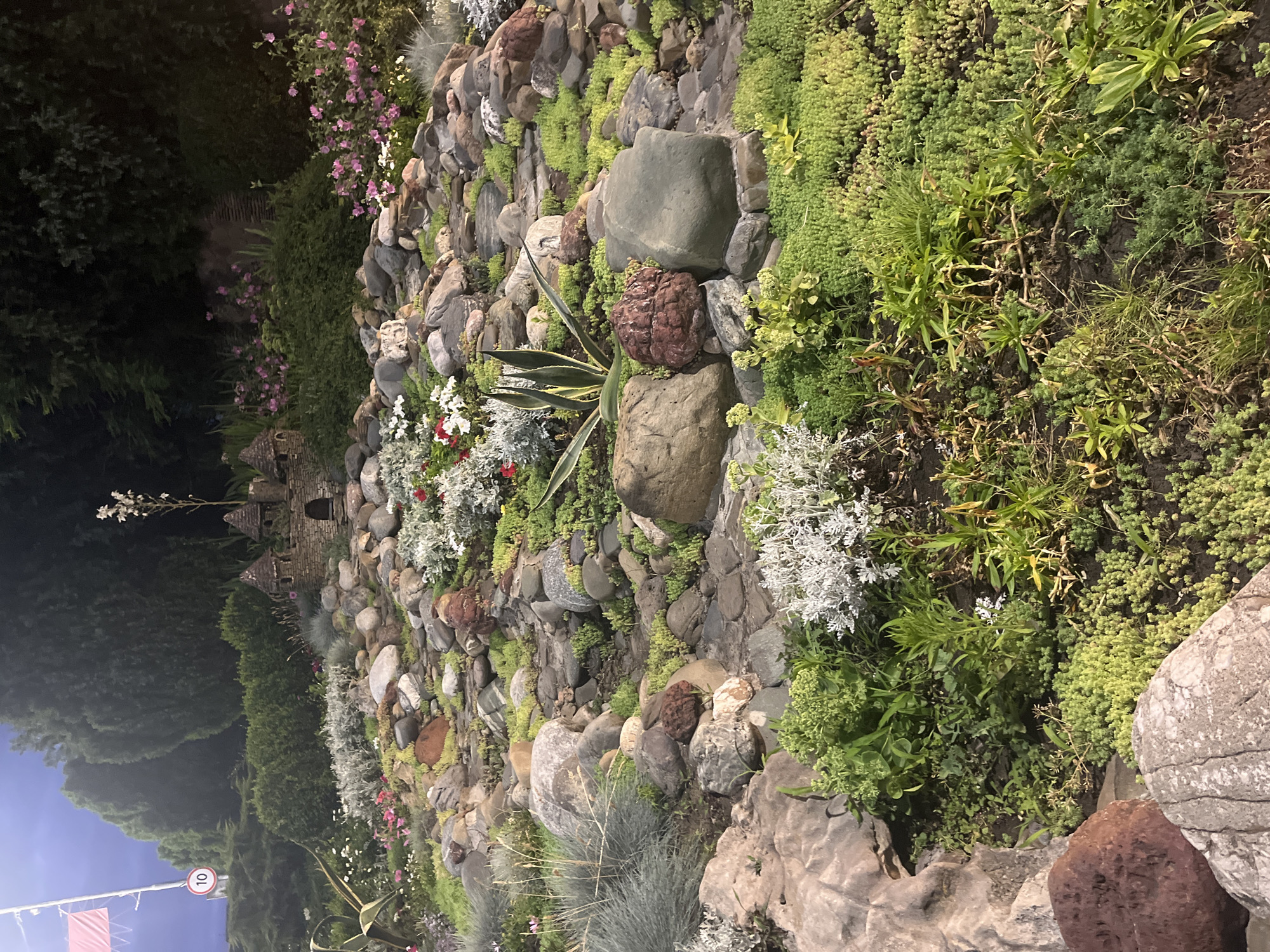 The image shows a miniature castle made of stones, located in a garden area. The castle is surrounded by rocks and various plants, including succulents, low-lying ground cover, and some flowering plants. In the background, there are trees and bushes. A street sign with the number "10" is visible on the left side of the image, along with a red and white flag. The lighting suggests it is either dusk or an artificially lit environment. The scene is situated in a garden in the city of Krasnodar, Russia.