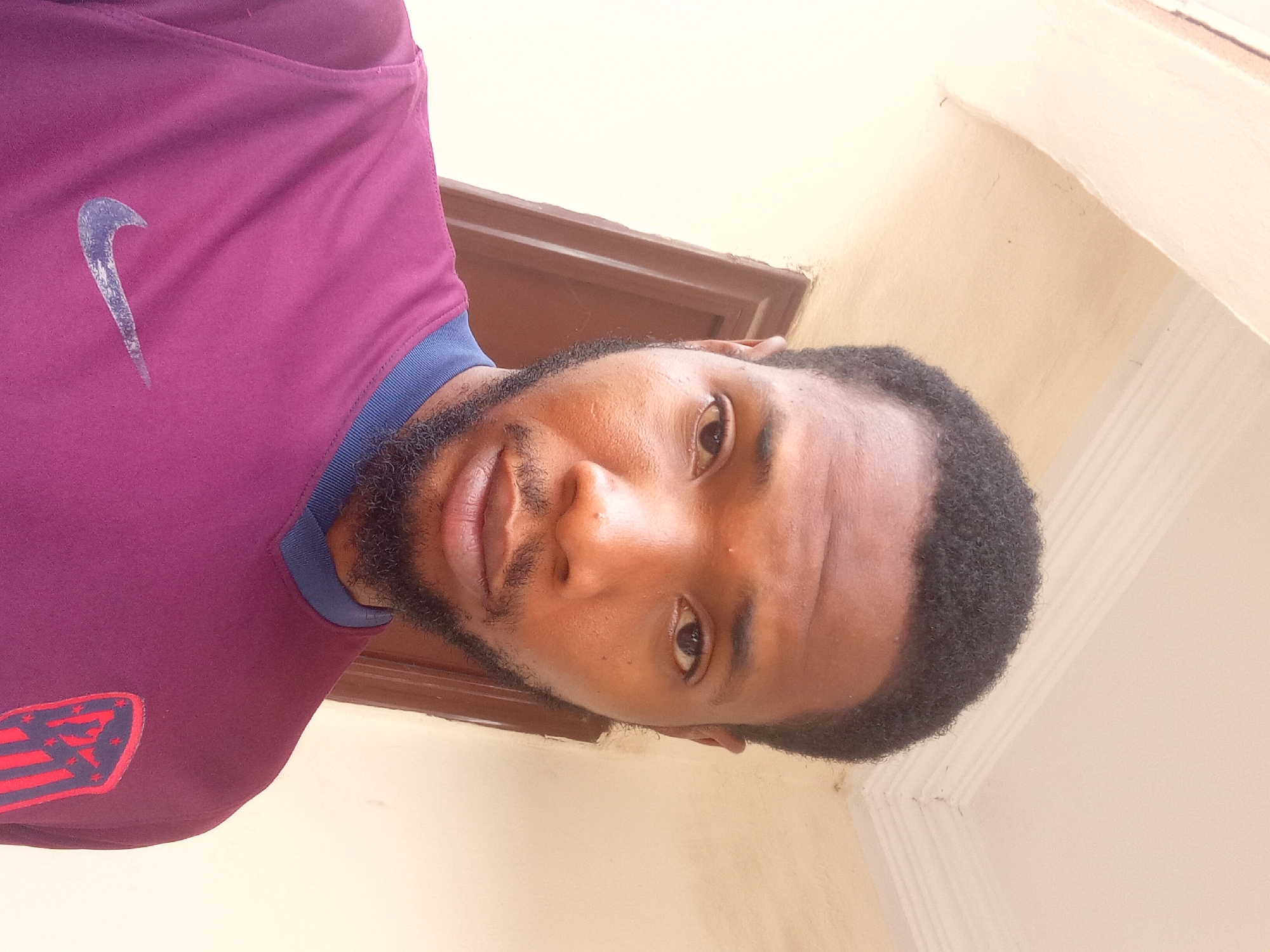 The image shows a man with dark hair and a beard wearing a maroon Nike athletic shirt with the Atletico Madrid logo on the left chest. He appears to be indoors, standing in front of a cream-colored wall and brown trim. The location is likely Zango-ndaji, Nigeria.