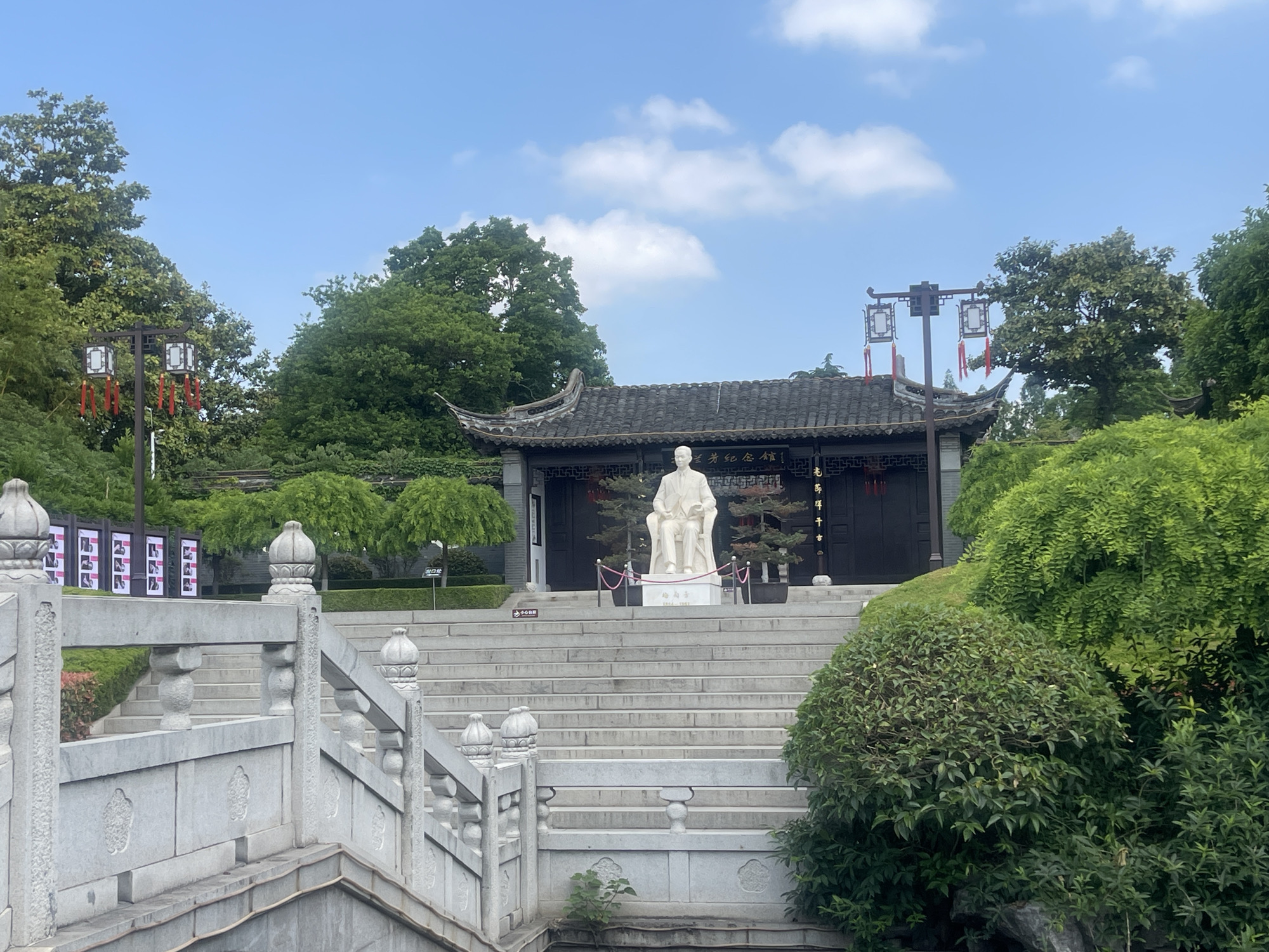 The image depicts a statue in front of a building in Hailing District, China. The statue, a light-colored figure seated in a chair, sits atop a series of concrete steps leading up to a dark-roofed building with traditional Chinese architectural elements. The building is decorated with hanging lanterns and framed by trees and greenery on either side. A stone bridge with ornate railings is in the foreground, partly obscured by foliage. The sky above is blue with scattered clouds.