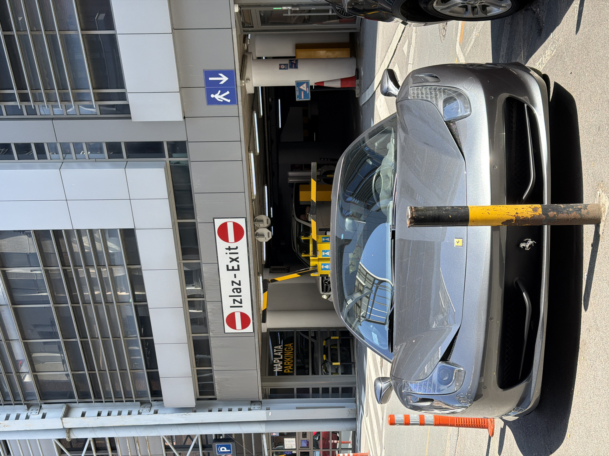 A grey Ferrari sports car, likely a 458 Italia model, is parked in the foreground on an asphalt surface, facing directly towards the viewer. A black and yellow striped bollard stands immediately in front of its bumper. To the left of the Ferrari, an orange and white striped traffic bollard is visible. A dark-colored SUV is partially visible to the right of the Ferrari.
In the midground, behind the Ferrari, is the entrance to an underground parking garage. Above the main vehicle exit, a prominent white sign with red borders displays "Izlaz-Exit" (Serbian and English for "Exit") flanked by two red "No Entry" symbols. To the left of the exit, above a pedestrian entrance, a yellow sign with black text reads "NAPLATA PARKINGA" (Serbian for "Parking Payment"). A yellow and black striped barrier arm is visible in a raised position within the vehicle exit. To the right of the vehicle exit, above a pedestrian walkway, a blue square sign with a white walking person icon and a downward arrow indicates a pedestrian route. Inside the parking structure, a red and white traffic cone is visible. To the far left, a blue square sign with a white "P" and an arrow points to the left, indicating parking.
The background features a multi-story, modern building with a facade characterized by horizontal metallic louvers and large glass windows reflecting the sky and surrounding structures. The building's structure includes visible grey support columns and paneling. The scene is illuminated by bright daylight, casting distinct shadows. The location is the Garaža Vukov Spomenik (Vuk's Monument Garage) in the Vračar Urban Municipality, Belgrade, Serbia. No people are visible in the image.