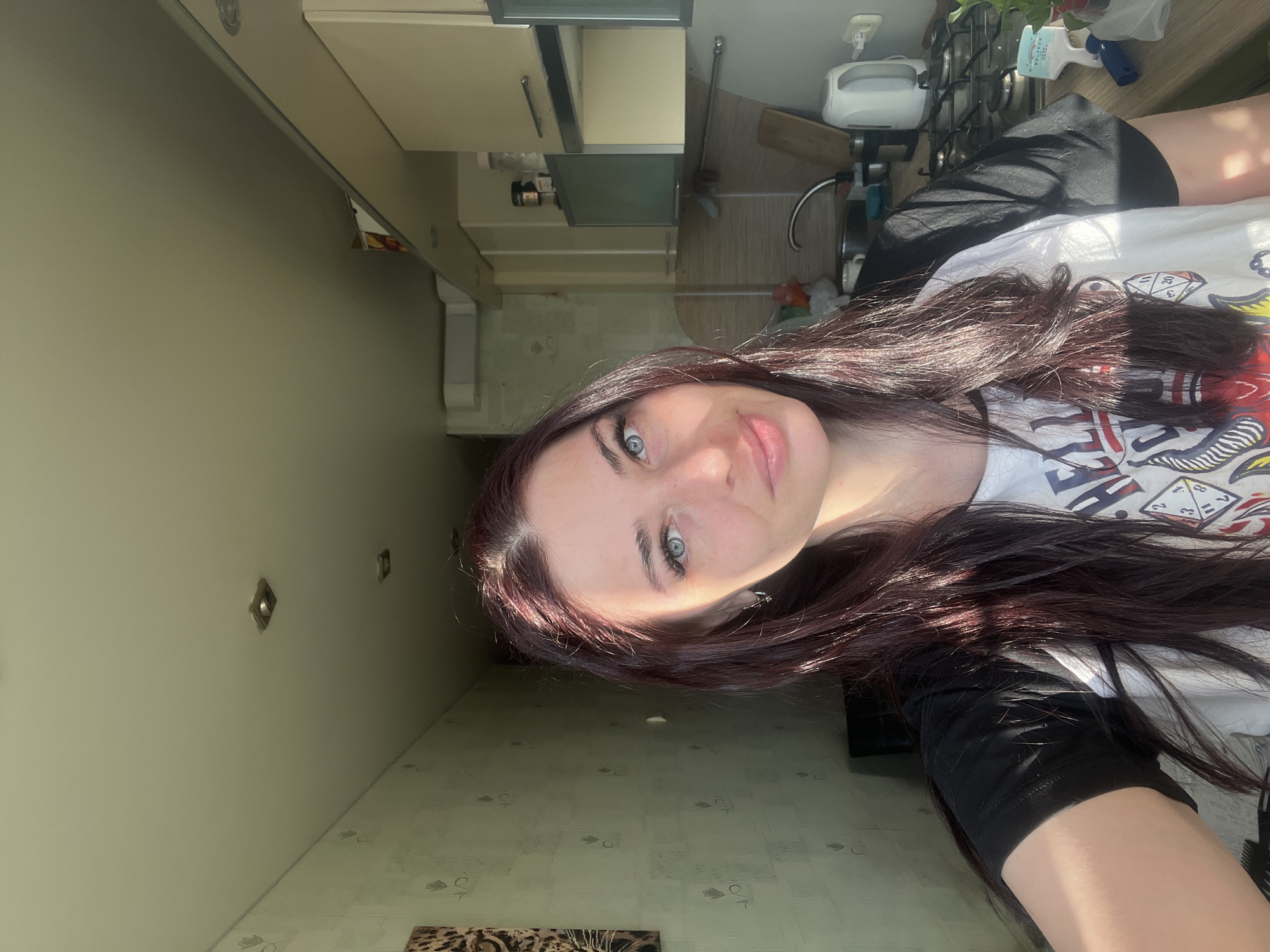 The image is a selfie of a woman with long, auburn hair and blue eyes, possibly taken indoors. She is wearing a white t-shirt with a black sleeve and black shorts. The background shows what appears to be a kitchen or dining area with light-colored walls and cabinets. There is a table or countertop visible, as well as what could be kitchen appliances. The floor has a patterned design. The image was taken at an angle, giving a perspective of the room behind and to the side of the woman. The location provided, Chaikovsky, Russia, is likely where the photograph was taken.