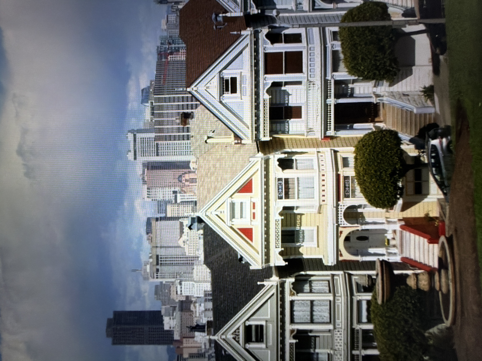 The image displays the Painted Ladies Victorian houses in San Francisco, California. The buildings are depicted with a cityscape in the background under a sky with clouds. The foreground shows the facades of the houses with details like windows, eaves, and foliage. Cars are parked on the street in front of the houses. The image is presented rotated 90 degrees counter-clockwise. Pétange, Luxembourg is not depicted in the image.