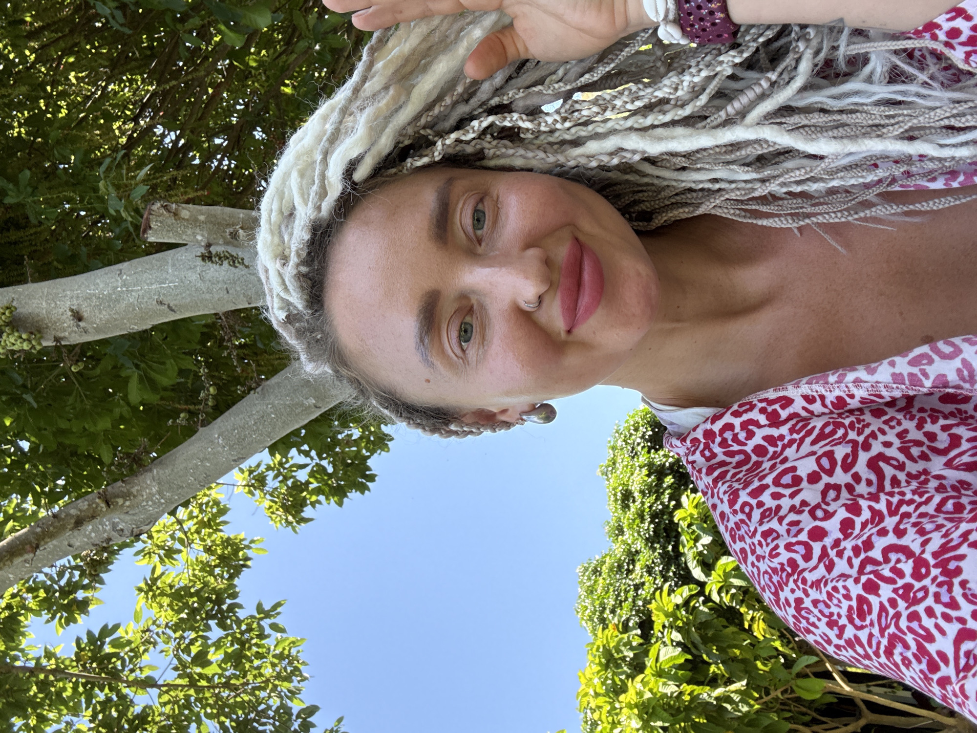 In a close-up selfie, a woman with pale skin and long, white braided hair poses outdoors against a backdrop of lush green foliage and a clear blue sky. She is wearing a white top with a vibrant red and white leopard print overlay. Her left hand is raised, partially obscuring her face, and her right hand is visible near her chest. She has blue eyes, a small nose ring, and is wearing large, teardrop-shaped earrings. Her lips are colored with a matte pink lipstick. The image appears to have been taken on a sunny day, with the sunlight highlighting her skin and hair. The environment suggests a tropical or garden setting. The specific location in the city of Bac Nha Trang Ward, Vietnam, is not visually evident in the image.