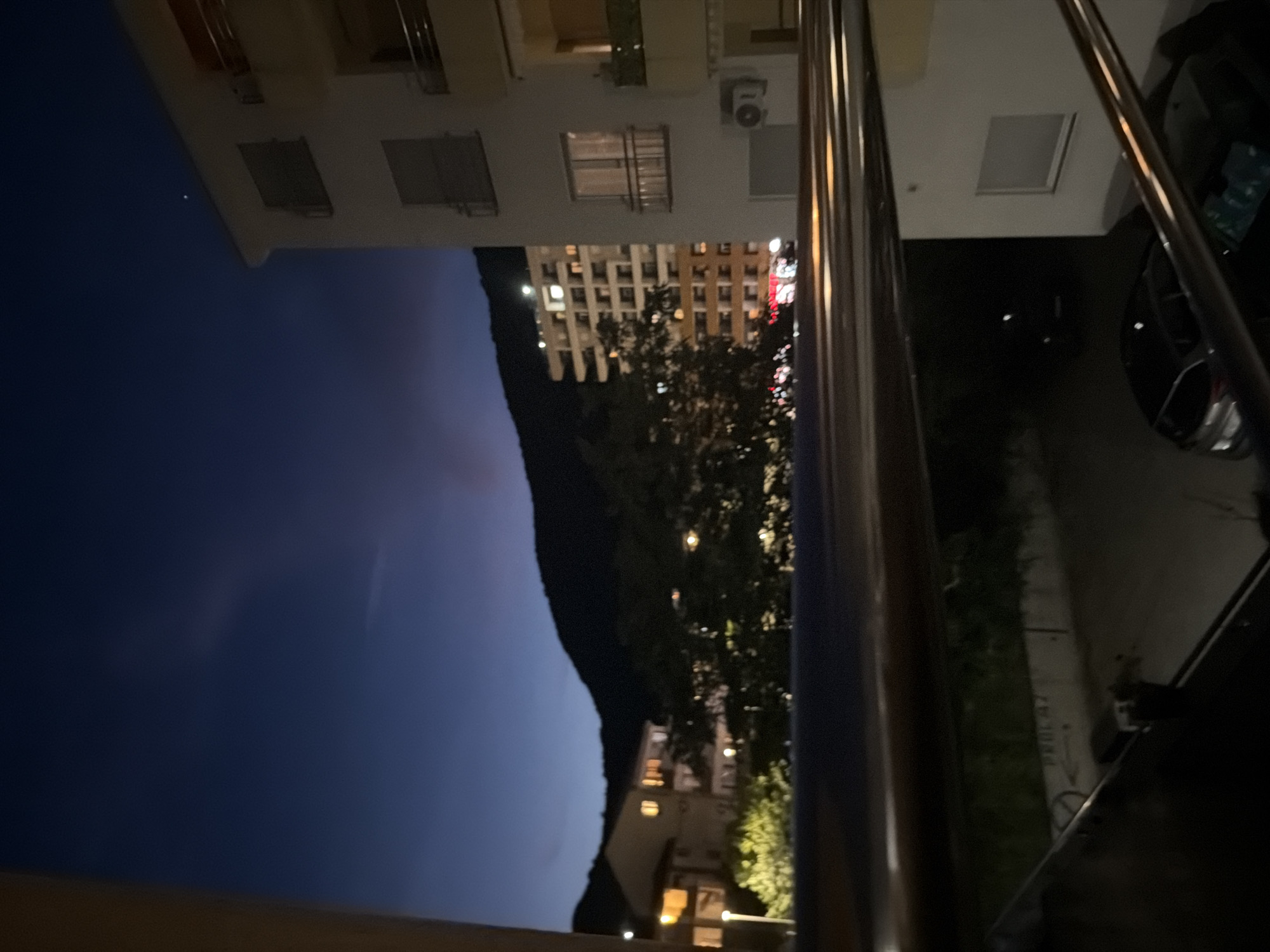 A nighttime view from a balcony in Budva, Montenegro, looking out over buildings and a mountain. The sky is a deep blue, transitioning to a lighter hue near the horizon. On the right side of the image is a white apartment building with balconies and windows. To the left, another building with illuminated windows is visible, nestled against the dark silhouette of a mountain. Trees are scattered throughout the scene. In the foreground, a metal railing runs horizontally across the image. Below the railing, a street is visible with parked cars and streetlights.