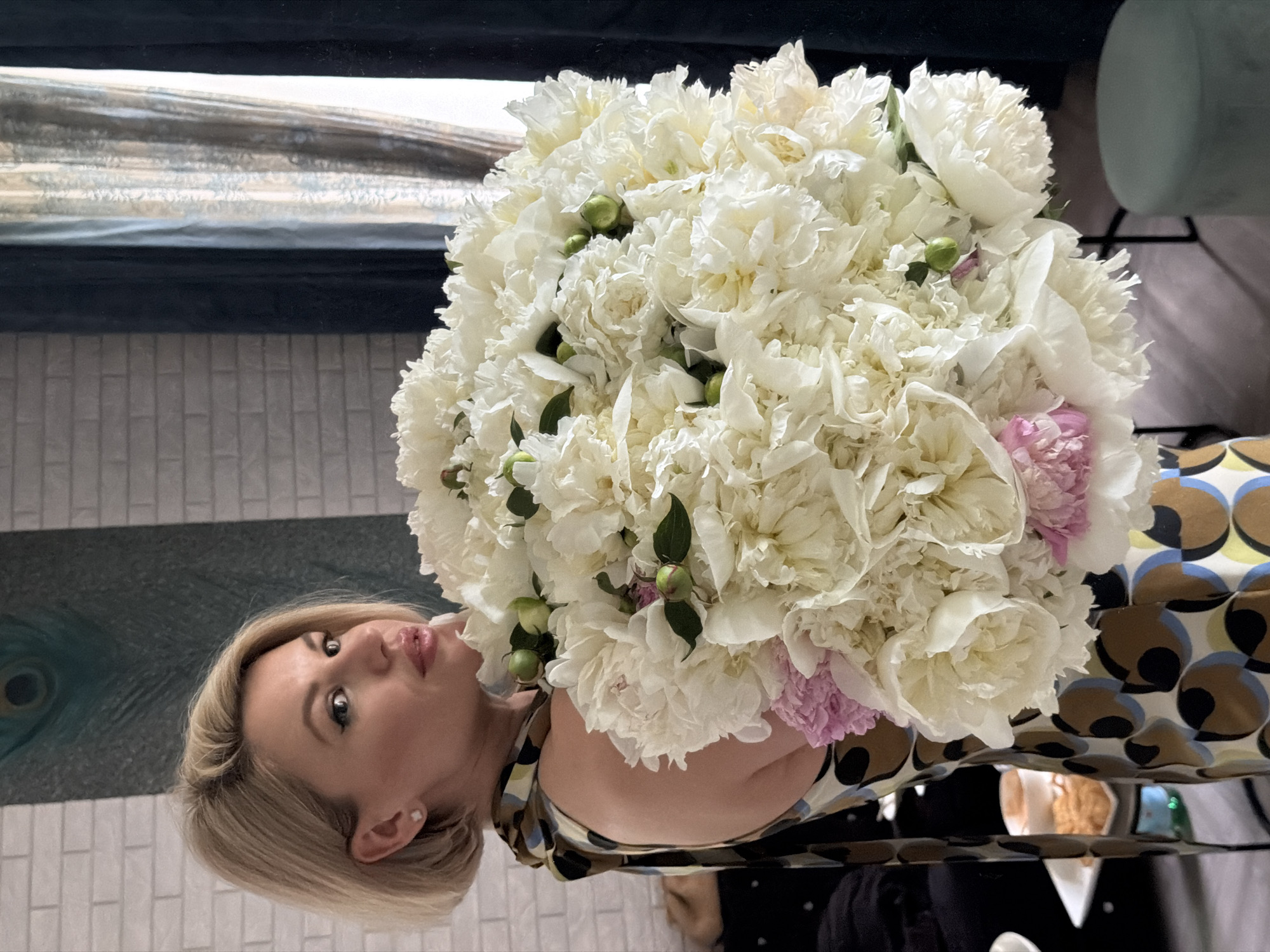 A blonde woman wearing a sleeveless dress with a multicolored geometric pattern is holding a large bouquet of white peonies and a few pink peonies. She is standing indoors, with a brick wall and a painting of a peacock feather on the wall behind her. To her right, dark blue curtains are visible. The woman has on earrings and makeup, and she is looking to her left. In the foreground, on a table, is a plate with cookies and a cup of coffee. A green stool is partially visible in the lower right corner of the frame. There is no information about the location in Riga, Latvia.