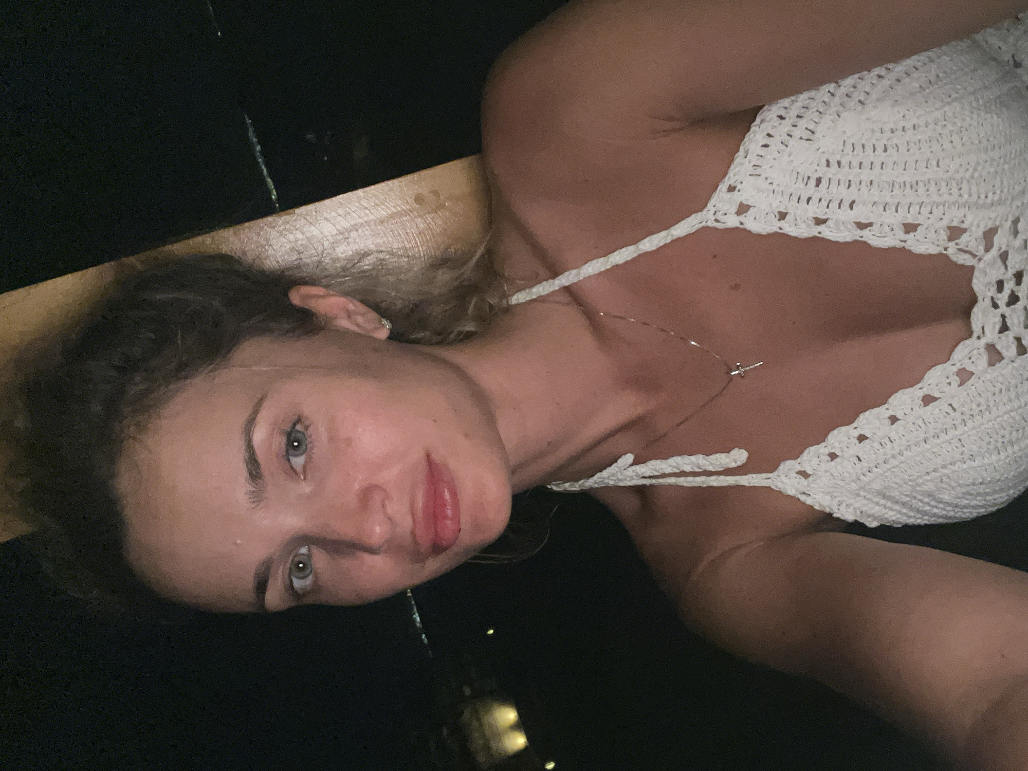 A woman with blonde hair pulled up in a bun is shown from the chest up. She is wearing a white crocheted halter top with a cross necklace. Her skin is tanned and her eyes are light blue. She is looking at the camera with a slight smile. The background is dark with some blurry lights in the distance, possibly indicating an outdoor setting at night. The woman appears to be in a resort or beach setting, potentially in the city of Nuweiba, Egypt, known for its beautiful coastlines and relaxed atmosphere.