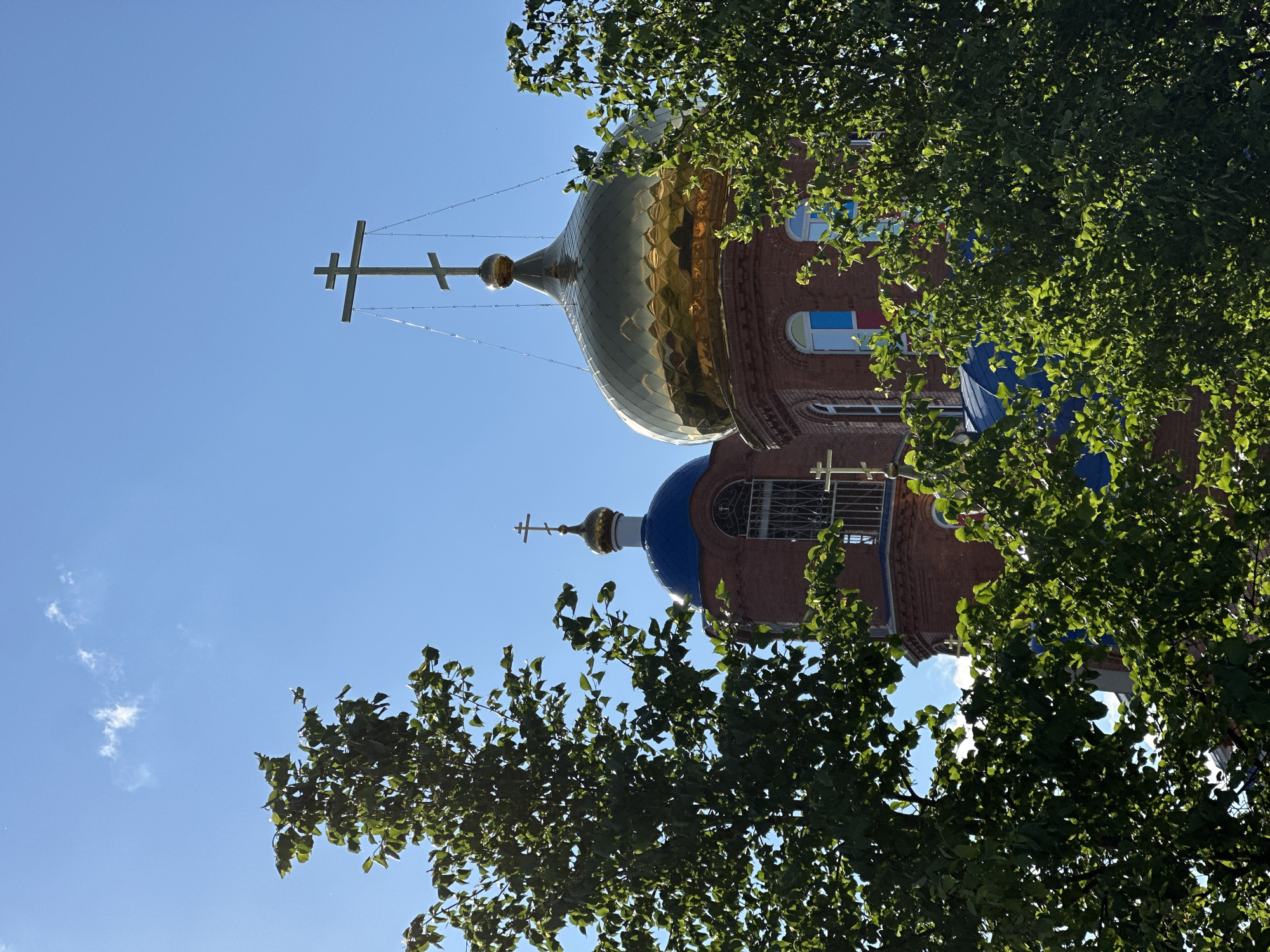 The image shows a brick church with golden and blue domes in Unknown, Unknown. A cross is perched atop each dome. The church is partially obscured by trees with lush green leaves. The sky is clear and blue with some faint white clouds visible.