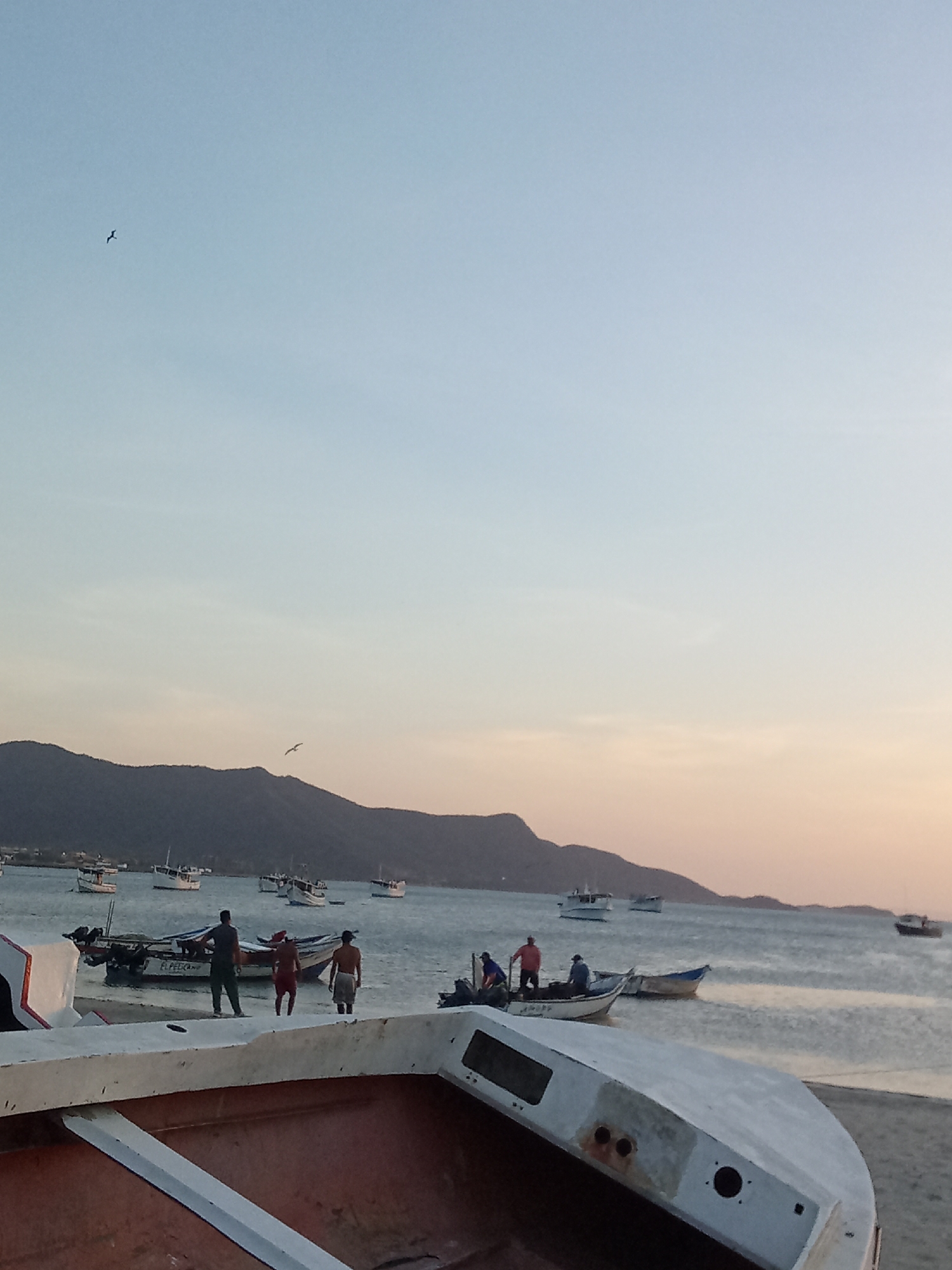 The image depicts a coastal scene in Juan Griego, Venezuela, at what appears to be dusk. The sky exhibits a gradient from light blue to a warm orange-pink hue near the horizon. A mountain range is silhouetted against the sunset, stretching across the middle of the frame.
The foreground is dominated by an overturned boat, its hull revealing a reddish-brown interior. In the midground, several boats are visible both on the shore and in the water. Some people are standing near the boats, seemingly preparing for or returning from a trip. One man is wearing a shirt and dark pants. Another is shirtless with red shorts, and a third man is shirtless wearing a white shorts.
The water is calm, with gentle ripples visible. A few additional boats are further out at sea. A single bird is soaring in the upper left of the frame.