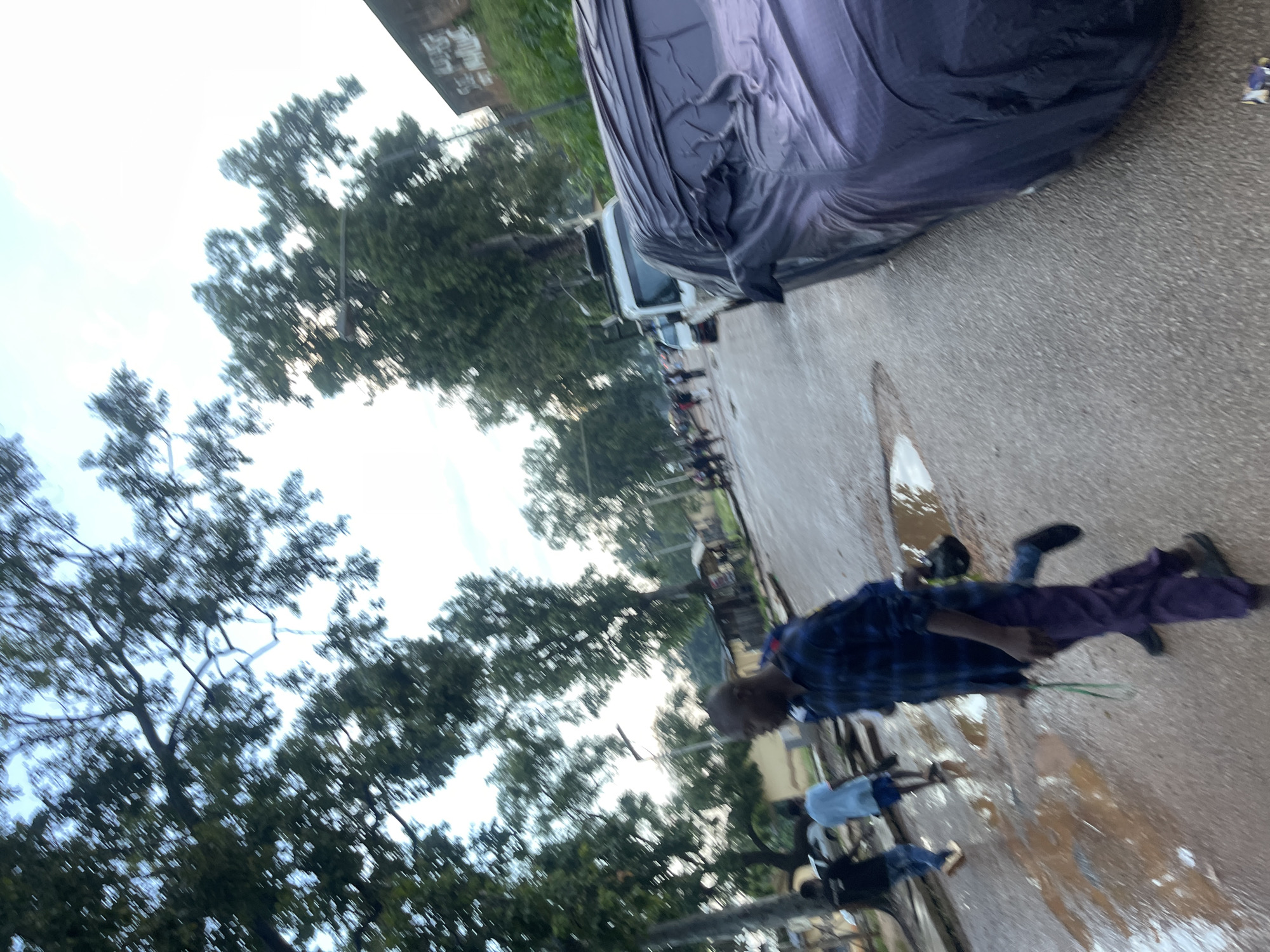 The image depicts a street scene in Rafin Par, Nigeria. In the foreground, a man wearing a blue and black checkered shirt and purple pants is walking to the right, holding a green string in his left hand. To his left, a man in a dark shirt and blue jeans is walking away from the camera. Further down the street, another person in a light blue shirt and blue shorts is walking to the right. A car covered with a dark purple tarp is parked on the right side of the road. The street surface is wet and shows several puddles, suggesting recent rainfall. Trees line the street, and some buildings are visible in the background. The sky is overcast.