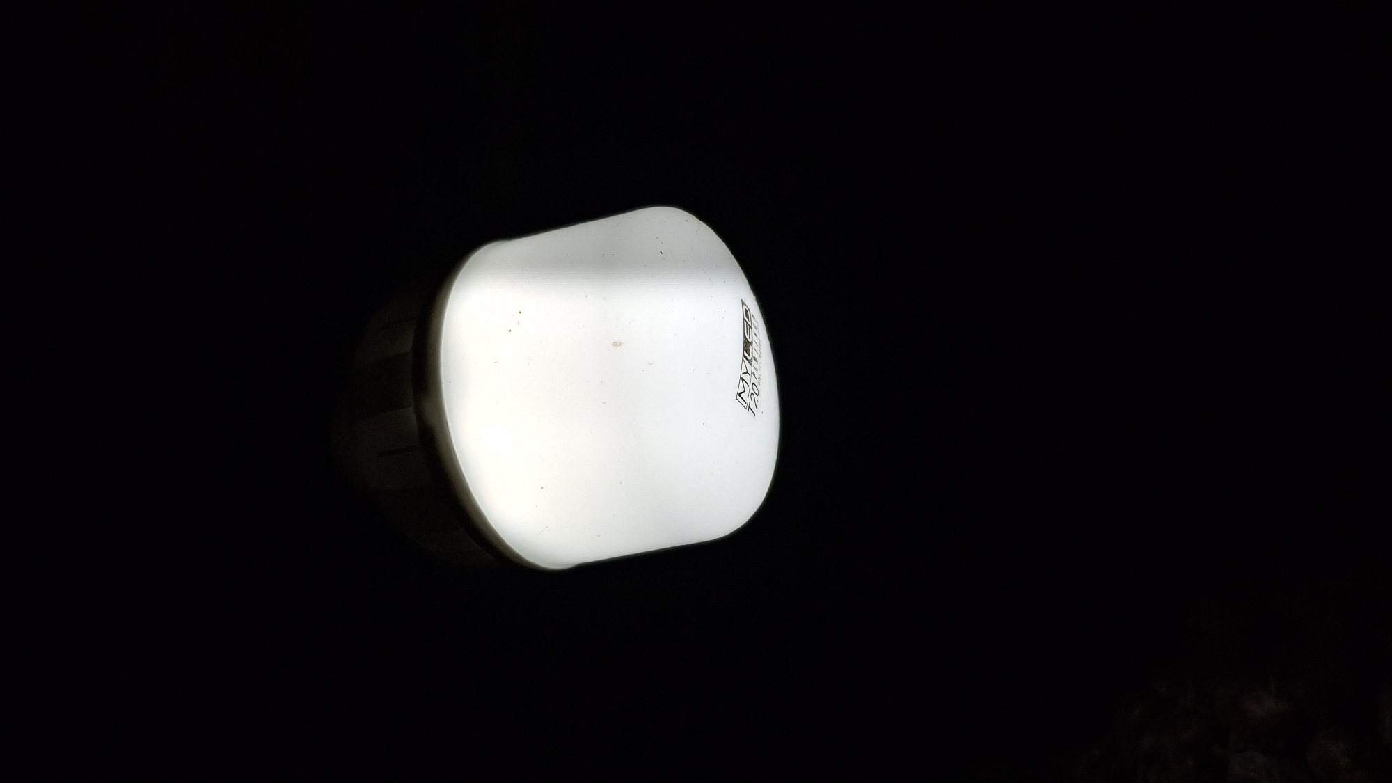 A close-up, low-angle shot shows a white LED light bulb against a black background. The bulb is lit and emits a bright white light. The brand name "MYLED" and the number "120V" are visible printed on the bulb in faint gray text. The image was taken in Pucakwangi, Indonesia. The scene is dark and featureless, except for the illuminated bulb.