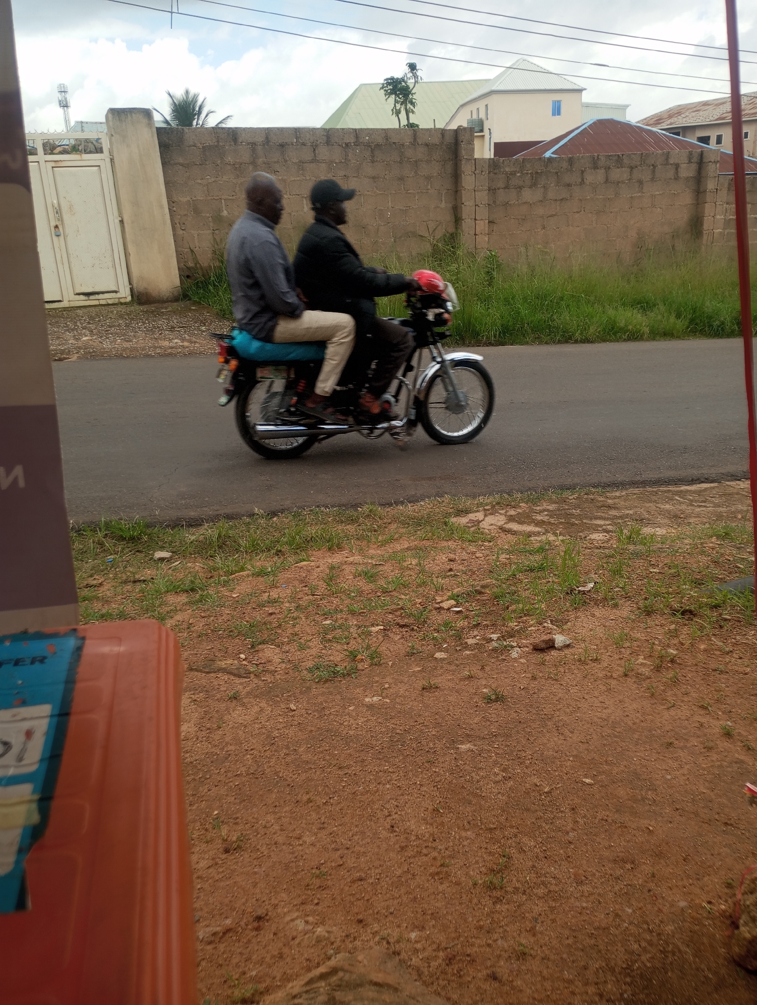 Two men are riding a motorcycle on a paved road in front of a brick wall. The man in the front is wearing a grey shirt and light-colored pants, while the man behind him is wearing a black jacket and a red helmet. The motorcycle has a blue seat. In the background, there are buildings and trees. The scene appears to be in Jos, Nigeria, based on the visual context.