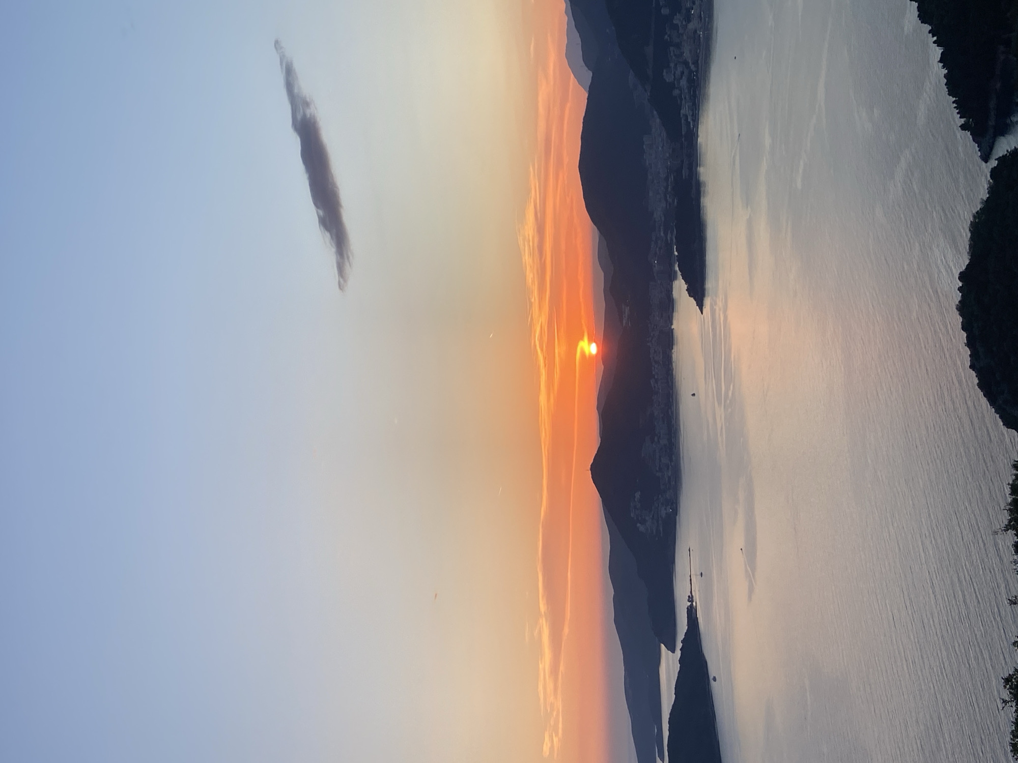 The image presents a landscape view with a sunset. To the left is a light blue sky transitioning to a yellow-orange horizon where the sun is setting behind mountains. A single cloud is in the upper left corner. A body of water, likely the Adriatic Sea, stretches across the right side of the image. Sveti Stefan is likely located along the mountain range on the right side of the water. Vegetation appears to be at the edge of the water on the right side of the image. Boats are on the water and buildings on the hillside.
