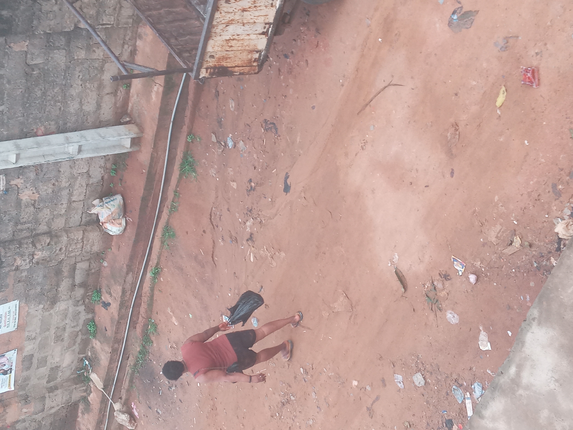 The image depicts a scene in Nkpor, Nigeria from an elevated perspective. The ground is primarily red-brown dirt, scattered with trash. A man is walking on the dirt; he is wearing a reddish-brown tank top and dark shorts and sandals, and carrying a dark bag. Part of a rusty metal structure is visible in the upper left quadrant of the image. Concrete walls are on the left and the bottom right. A small poster is on the left wall.