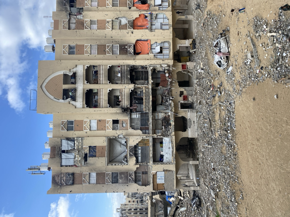 The image displays a multi-story building, appearing to be residential or mixed-use, severely damaged and standing amidst a vast field of rubble under a clear blue sky with scattered white clouds. The building, constructed from light-colored materials, shows extensive structural damage across multiple floors. Large sections of its facade are blown out, revealing gutted interiors with exposed concrete, twisted rebar, and remnants of household items. Many windows are shattered or missing, and some areas show burn marks and charring. On the upper levels, several large, bright orange tarp-like materials are visible within damaged sections. A distinctive arched, ornate feature is present on the upper part of the building's facade. White water tanks are visible on the roof or upper left side.
At the base of the building, a massive debris field extends outwards, comprising shattered concrete, metal fragments, and various building materials mixed with sand or earth. This rubble covers a significant portion of the ground. Towards the right side of the image, the ground transitions to sandy earth with fewer, smaller pieces of debris. Another partially visible building, appearing less damaged, can be seen in the background on the far left.
The scene captures the aftermath of a devastating event, likely an airstrike or heavy shelling, given the scale of destruction. The time of day is clearly daytime, indicated by the bright sky. No human figures or active events are discernible. No visible text is present. The overall scene conveys widespread destruction and the profound impact of conflict in an urban environment, consistent with the provided location context of Khan Yunis, Palestinian Territory.