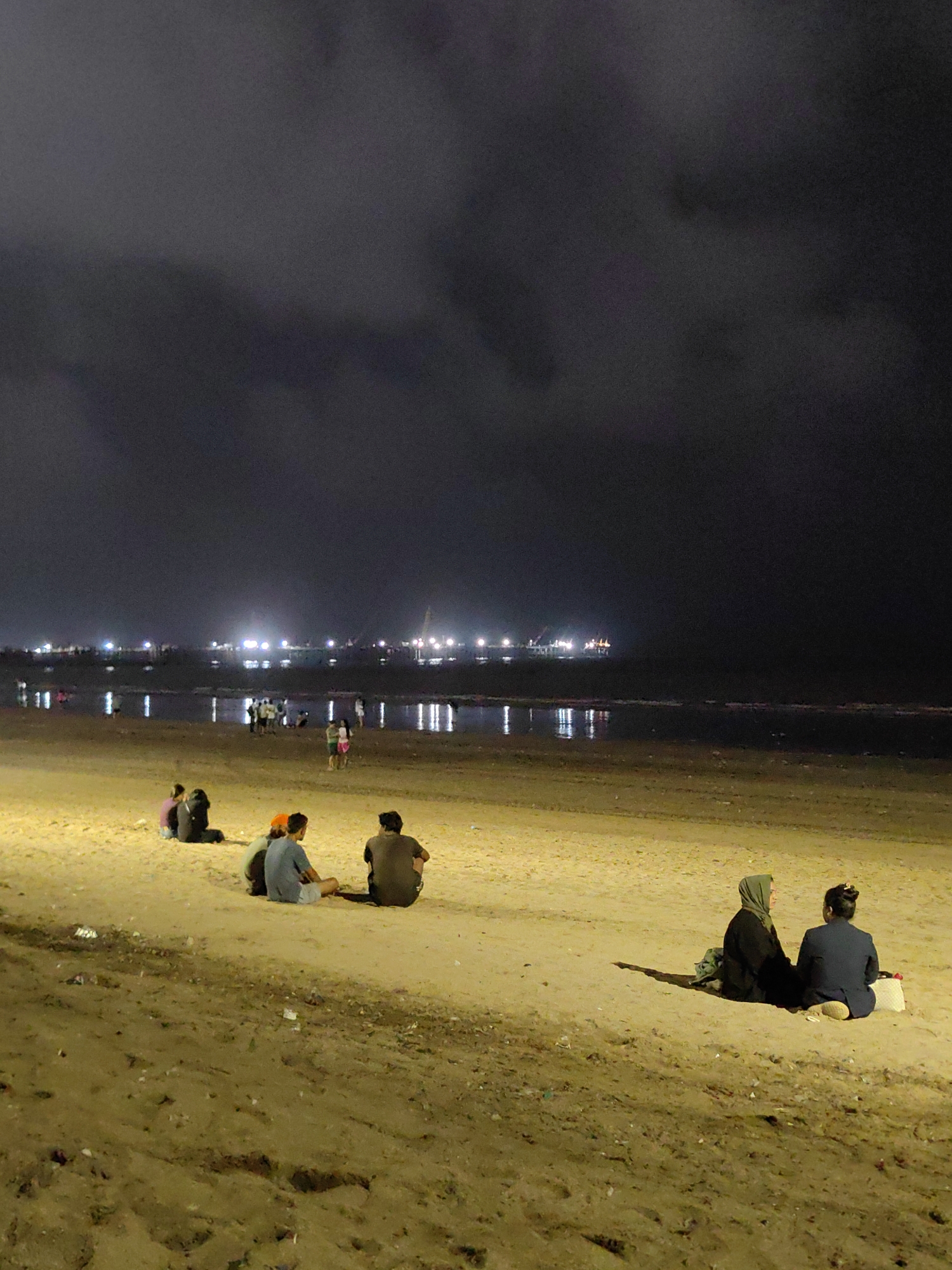 At night on Juhu Beach in Mumbai, India, several groups of people are sitting on the sand. In the foreground, on the right, two women are seated. One woman on the left is wearing a green hijab and a dark robe, and the woman on the right is wearing a dark blue blazer and a light-colored headscarf tied in a bun. To the left of them, four people are seated: two men in the center and two women further back on the left. The group on the left consists of two men, one wearing a blue shirt and the other wearing a brown shirt, with a person in between them wearing an orange hat. The two women on the far left are sitting with their backs to the camera.
In the midground of the image, there are several more people walking and standing near the water's edge. The sea is calm, and the reflection of distant lights can be seen on the water. In the background, a long line of lights suggests a distant city skyline or port area, possibly indicating the presence of ships or industrial structures. The sky is dark and cloudy. The sand is a light brown color and appears to have some scattered litter.