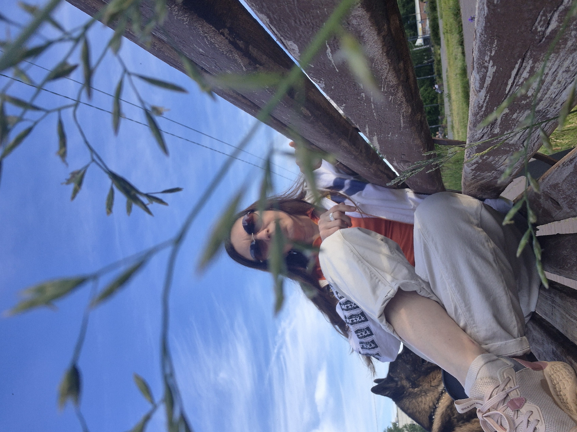 This low-angle image, taken in Chaikovsky, Russia, features a person sitting on a wooden bench outdoors under a bright blue sky. The main subject is a person, likely female, with long brown hair, wearing dark sunglasses, an orange top, and light-colored (possibly white or beige) pants. A white long-sleeved shirt with a repeating pattern of stylized text, appearing to read "AT SXE" or "SXSXE," is visible on the sleeve under the orange top. They are wearing light pink and beige sneakers. One hand is raised near their face, holding a small, indistinct object.
A dark-furred dog, possibly a German Shepherd or similar breed, is partially visible next to the person at the bottom right, its head and ear peeking into the frame. The background shows a vibrant blue sky with wispy white clouds, indicating clear daytime weather. Blurry green foliage occupies the foreground, framing the shot. In the distance, beyond the bench and trees, faint power lines and green open areas are visible. The scene suggests a relaxed outdoor setting, possibly a park or public area.