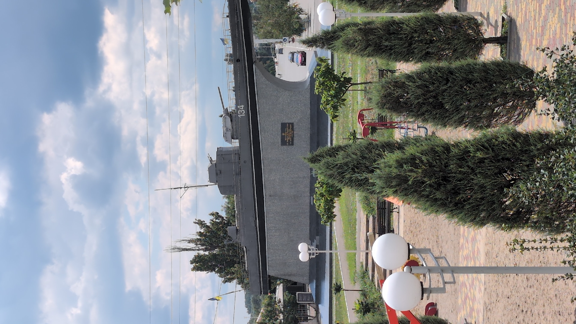 The image depicts a monument to a naval vessel, possibly a torpedo boat, in the city of Izmail, Ukraine. The monument is a large, dark grey vessel mounted on a grey, curved concrete pedestal. The vessel is oriented horizontally, with its bow facing left. It features visible weaponry, including a turret at the front and what appears to be a gun barrel extending from the middle. A number "134" is painted on the side of the vessel. A plaque is affixed to the pedestal, displaying text in a foreign language. A Ukrainian flag is visible to the left of the vessel. The monument is situated in an urban park area with paved walkways, benches, and landscaping, including evergreen trees and bushes. In the background, there is a road with a few vehicles, buildings, and trees. The sky is partly cloudy. In the foreground, a lamppost with two white spherical lights is visible, along with playground equipment and sand. The scene appears to be a public space commemorating a maritime event or figure associated with Izmail.