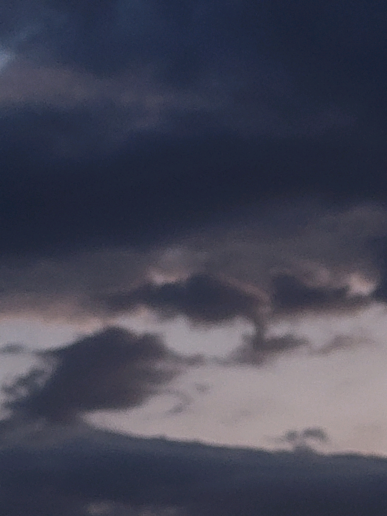 The image depicts a cloudy sky, likely at dusk or dawn, with dark, textured clouds dominating the frame. The lighting suggests a muted, atmospheric scene. There are no discernible people, subjects, objects, actions, or interactions visible. The environment is solely characterized by the cloud formations. The location in Ozolnieki, Latvia, is not visually represented in this image.