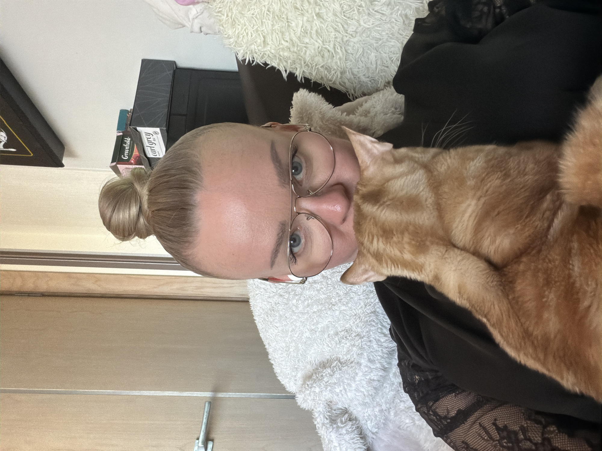 A woman with blonde hair in a bun and wearing round glasses is holding an orange tabby cat close to her face. The cat is nuzzling her. She is wearing a black top with lace detailing. There is a fuzzy white blanket or cushion behind her and other objects stacked behind her head including tea boxes that show the words "Greenfield" and "Earl Grey". A door or panelled wall is visible on the left side. The image was taken indoors and there is no information to confirm or deny its location in Murino, Russia.
