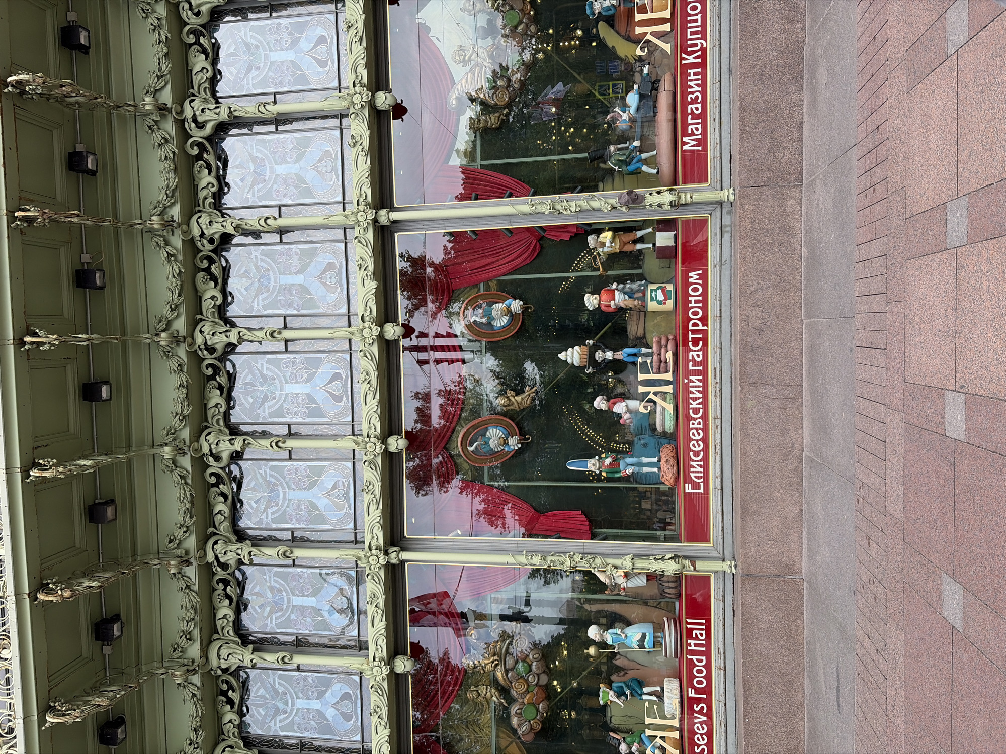 The image shows the exterior of the Eliseev Emporium, a historic food hall located on Nevsky Prospekt in Saint Petersburg, Russia. The building features an ornate Art Nouveau facade with large, decorative stained-glass windows. The storefront windows are adorned with elaborate displays of animated figures and festive decorations, reflecting the Emporium's reputation for extravagant window displays. Red velvet curtains are visible behind the figures, adding to the theatrical effect. The shop's signage is in both English ("Eliseev's Food Hall") and Russian ("Елисеевский гастроном" and "Магазин Купцов Елисеевых"). The pavement in front of the building is made of granite.