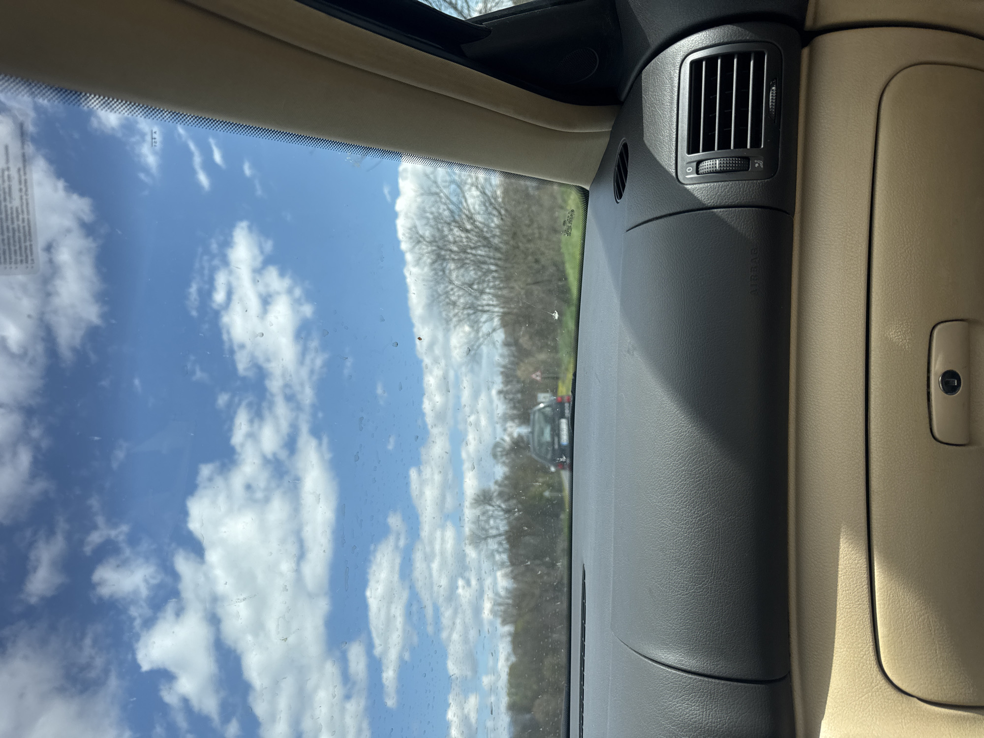 The image shows a view from inside a car, looking out through the windshield at a partly cloudy sky and trees. Another car is visible in the distance. The dashboard of the car is visible, including an air vent and glove compartment. The location is likely not Osetenovo, Bulgaria as it is impossible to determine the location from the image.