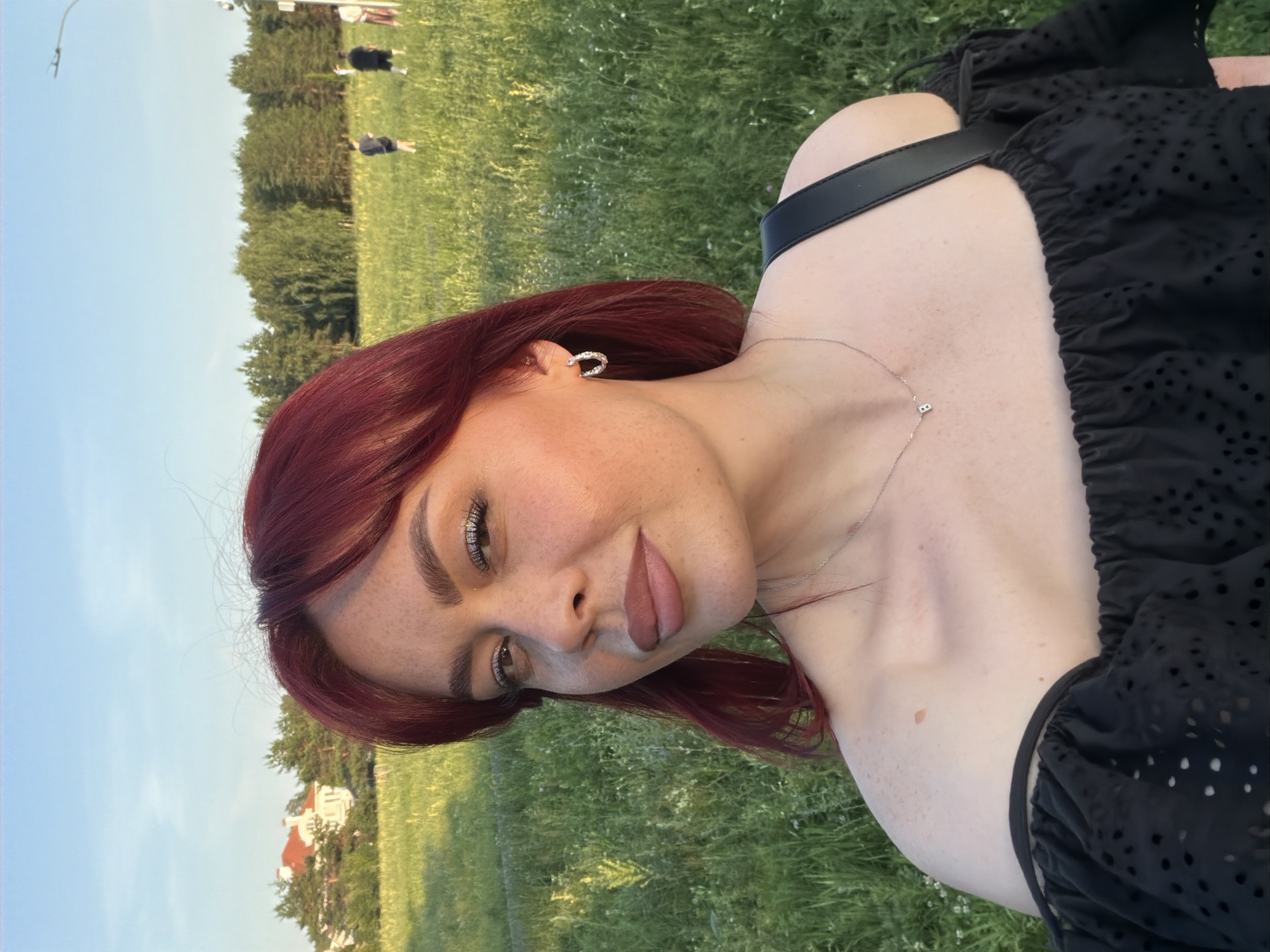 A young woman with red hair is captured in a selfie taken outdoors during daylight. She is positioned in the foreground, facing the camera with a slight smile. Her attire includes an off-the-shoulder black top with intricate cutouts. She is wearing silver hoop earrings and a delicate silver necklace with a small pendant. The background suggests a park or open field with lush green grass and scattered wildflowers. In the distance, a building with a red roof and trees are visible. A few individuals can be seen further in the background, engaged in recreational activities. The sky is clear and blue. This scene is consistent with locations found within the city of Tolyatti, Russia, specifically areas like Victory Park or the adjacent green spaces known for their recreational appeal.