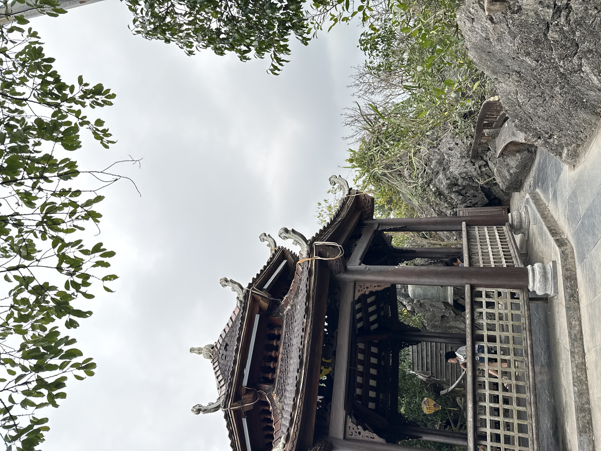 The image shows a low-angle view of a wooden pagoda structure with ornate roof detailing in Hoa Lư Ward, Vietnam. The pagoda is located near a rock face. The sky is overcast. There are trees in the foreground. People are seen inside the structure and walking up steps.