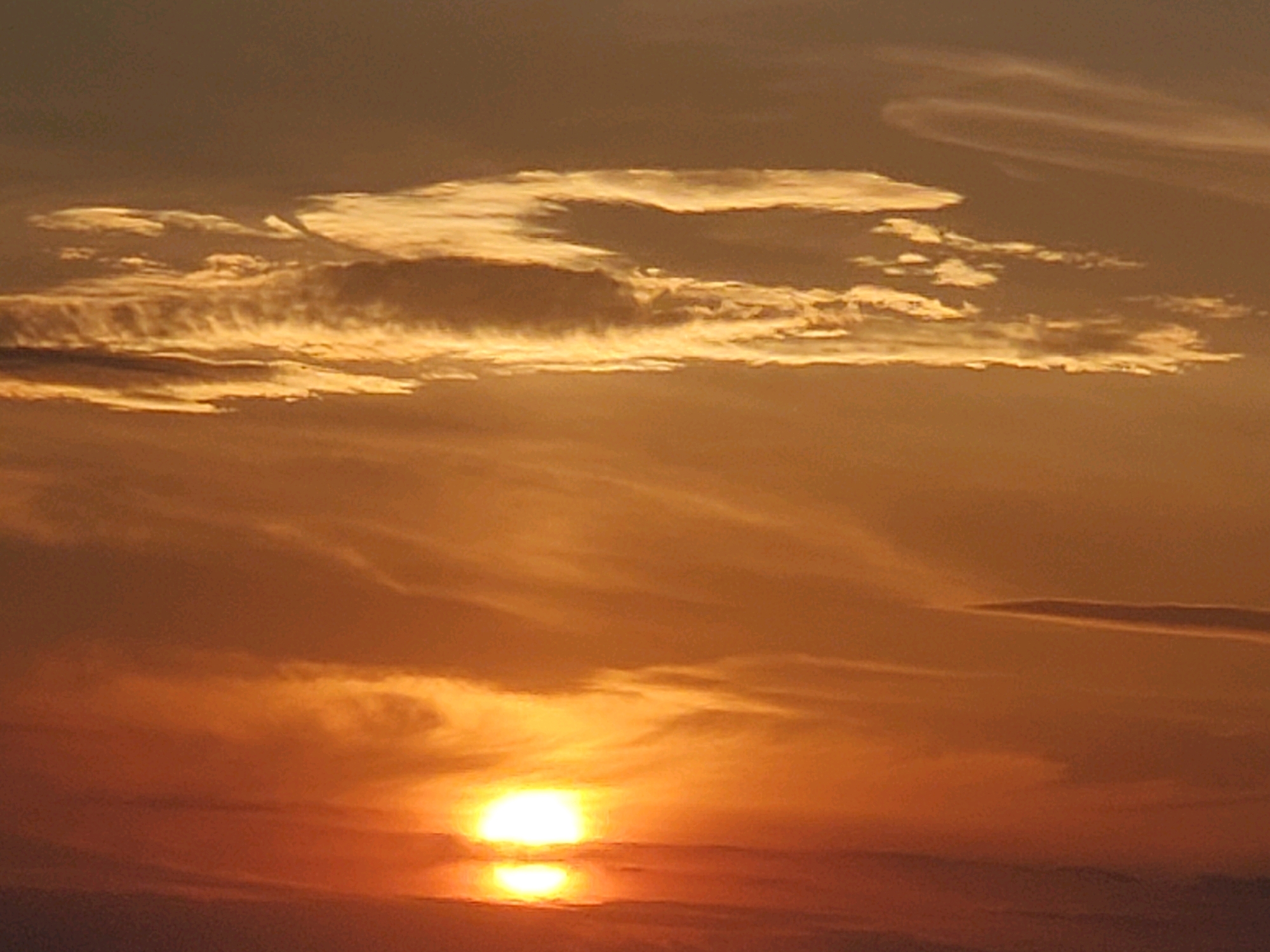 The image depicts a sunset over a body of water, likely a river or lake, in the city of Konstantinovka, Moldova. The sun, a bright, glowing orb, is partially obscured by clouds and sits low on the horizon, casting a warm, orange and yellow light across the sky and water. The clouds are wispy and streaky, catching the sunlight and creating a dramatic effect. The water reflects the colors of the sunset, creating a mirror image of the sky. The overall scene is serene and tranquil, with no visible people or objects to distract from the natural beauty of the sunset. The foreground appears to be a dark, indistinct expanse, possibly land or a shoreline. The sky transitions from a deep orange at the horizon to a muted brown at the top of the frame, with lighter orange and yellow hues in between.