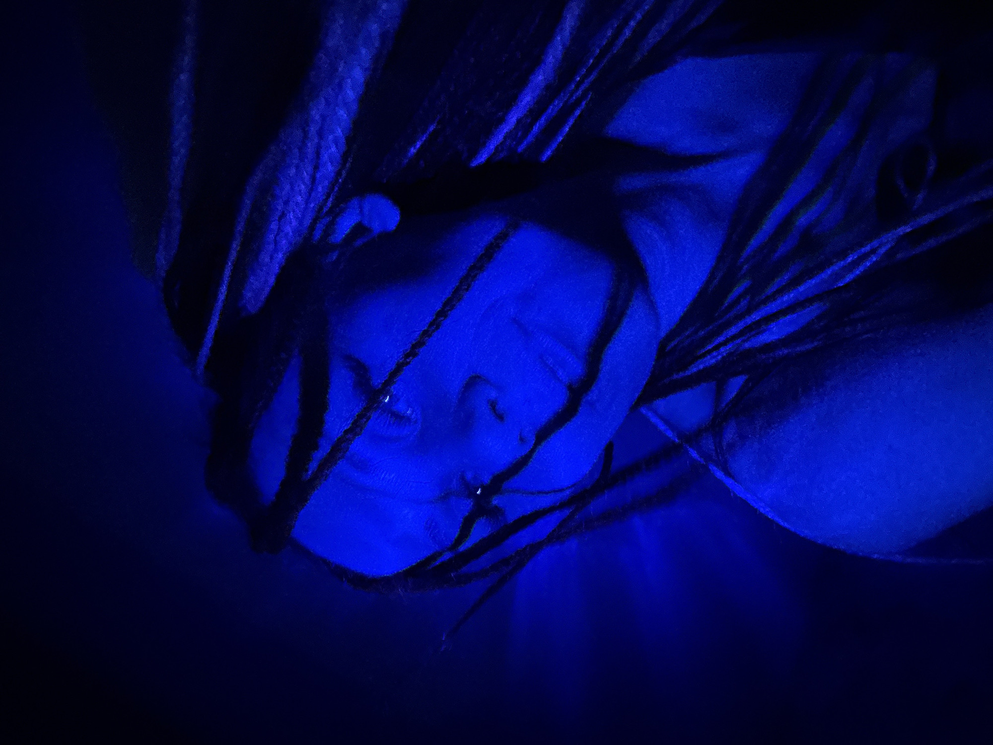A close-up image shows a person's face illuminated by a strong blue light. The person has braids or dreadlocks that frame their face and appear dark in the dim lighting. Their eyes are closed, and the overall impression is one of low visibility and strong color cast. The location cannot be identified from the provided image alone, nor can it be reliably determined that it is in Rusanovo, Russia. The information suggests that the lighting conditions consist of a dominant monochromatic blue light. No specific actions, interactions, or further environmental details are discernible from the provided image.
