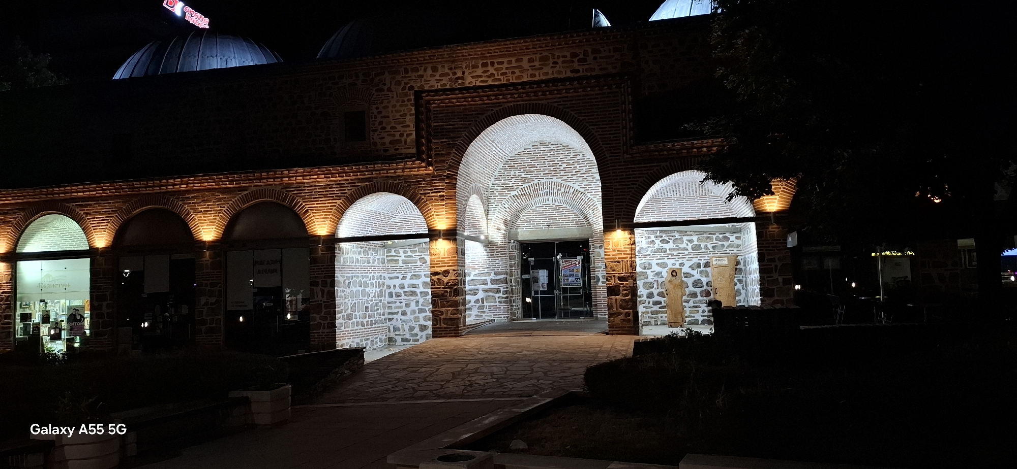 The image depicts the historic Kurşun Camii (mosque) in Yambol, Bulgaria, at night. The building is characterized by its arched entrances and traditional stone and brick construction. The facade is illuminated by warm lighting, highlighting the architectural details. Several shops or establishments are visible through the glass storefronts on the left side of the building. The foreground features a paved walkway and landscaping. The "Galaxy A55 5G" watermark is present in the lower left corner of the image.