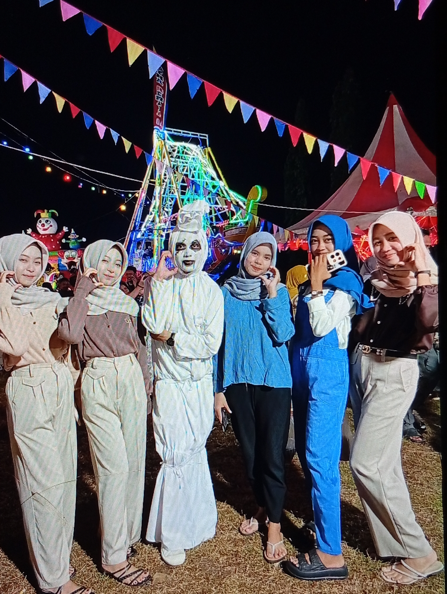 Six individuals, predominantly young women, are captured in a posed group photograph at an outdoor night fair in Lhokseumawe, Indonesia. The scene is illuminated by multi-colored lights from fair rides, string lights, and a prominent red-and-white striped carnival tent in the upper right background. Colorful triangular bunting flags are strung across the top of the frame. The ground consists of dry earth or sparse grass.
From left to right:
1. An individual wearing a light beige hijab, long-sleeved shirt, and wide-leg pants, smiling with their left hand near their face.
2. An individual wearing a light brown hijab, long-sleeved shirt, and light beige wide-leg pants, smiling and forming a heart shape with both hands.
3. An individual in a white, full-body ghost-like costume with white face paint and dark eye/mouth makeup, forming a heart shape with the adjacent individual.
4. An individual wearing a blue hijab, blue long-sleeved top, and black pants, smiling and forming a heart shape with their right hand, interacting with the costumed individual.
5. An individual wearing a blue hijab, white long-sleeved shirt, and blue denim overalls, smiling and holding a white-cased smartphone with their right hand near their face.
6. An individual wearing a light beige hijab, dark brown long-sleeved button-up shirt, light beige wide-leg pants, and a dark belt with a gold buckle, smiling with their right hand near their face.
Various sandals and flip-flops are worn by the individuals. A large, smiling clown or snowman figure is visible in the left background, behind the first two individuals.