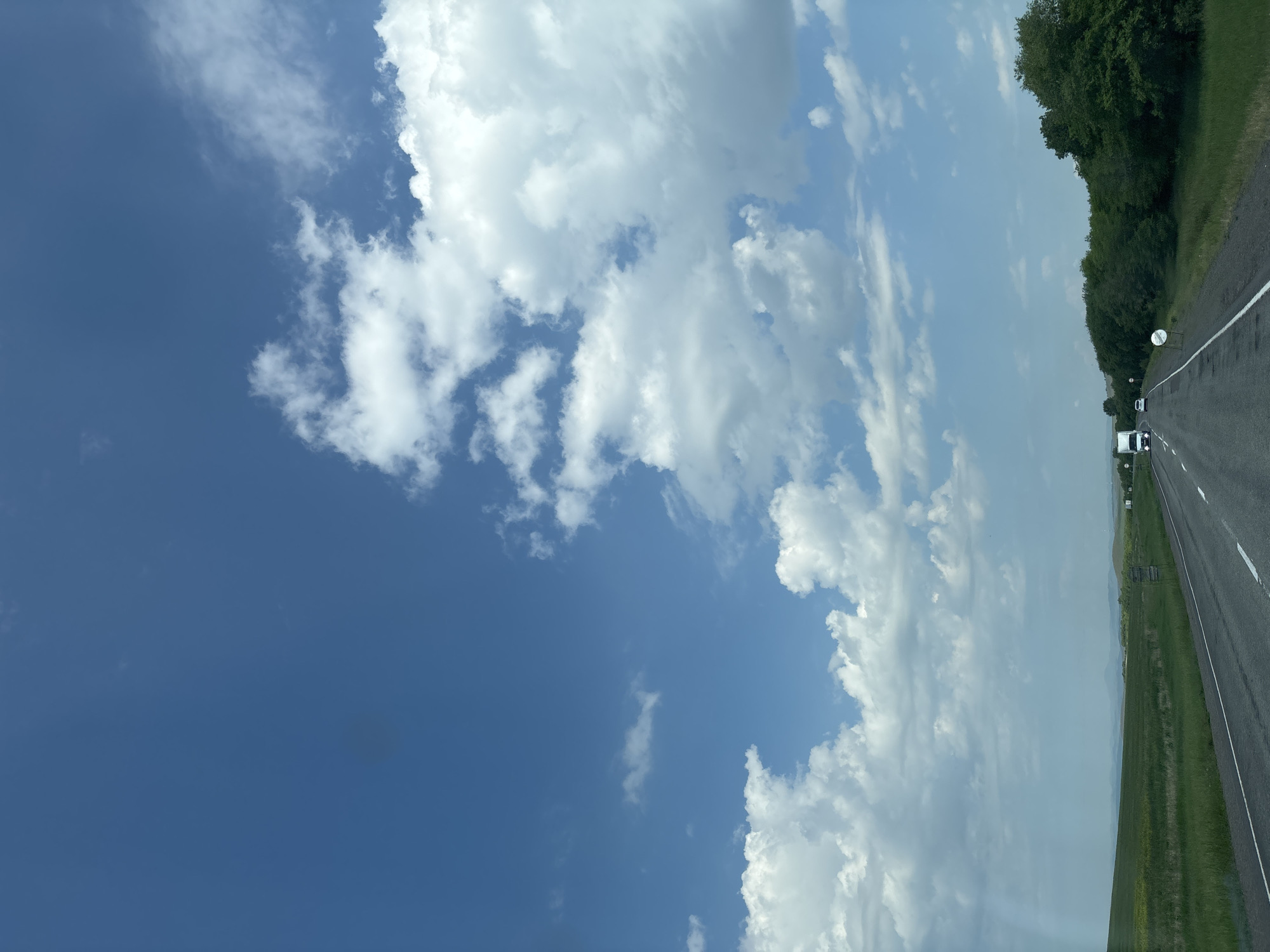 The image depicts a highway scene in the city of Borodino, Russia. A two-lane asphalt road stretches into the distance, bordered by green fields on the left and a dense line of trees on the right. The sky is a vibrant blue, dotted with fluffy white clouds. Several vehicles are visible on the road, including a white truck and a white car. The overall impression is one of a clear, sunny day on a rural highway.