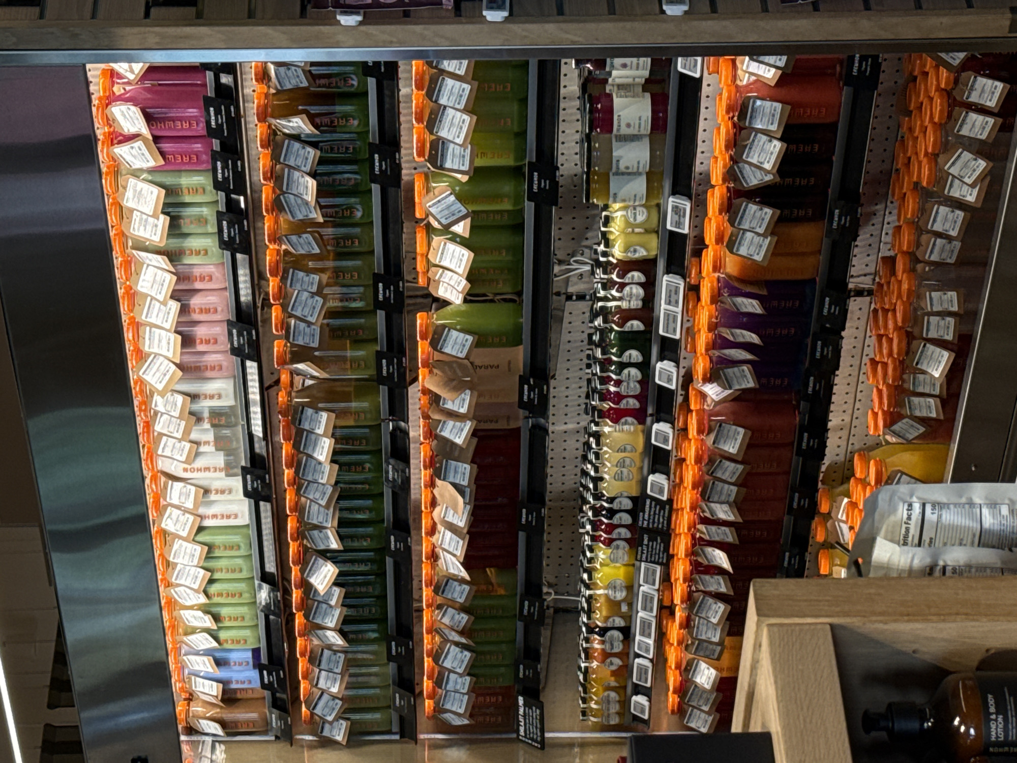 The image presents a refrigerated display case filled with a variety of Erewhon brand beverages, likely located within the Erewhon Market store in Beverly Hills, United States of America. The beverages are arranged in vertical columns, each column featuring various flavors and colors. Attached to each bottle is a label with branding and nutritional information. In the foreground, a wooden display shelf partially obscures the view of some of the refrigerated beverages. On the shelf, there is a brown bottle labeled "Hand & Body Lotion", with a white label marked "Nutrition Facts". The overall scene depicts a well-stocked, refrigerated beverage section within a high-end grocery store setting.