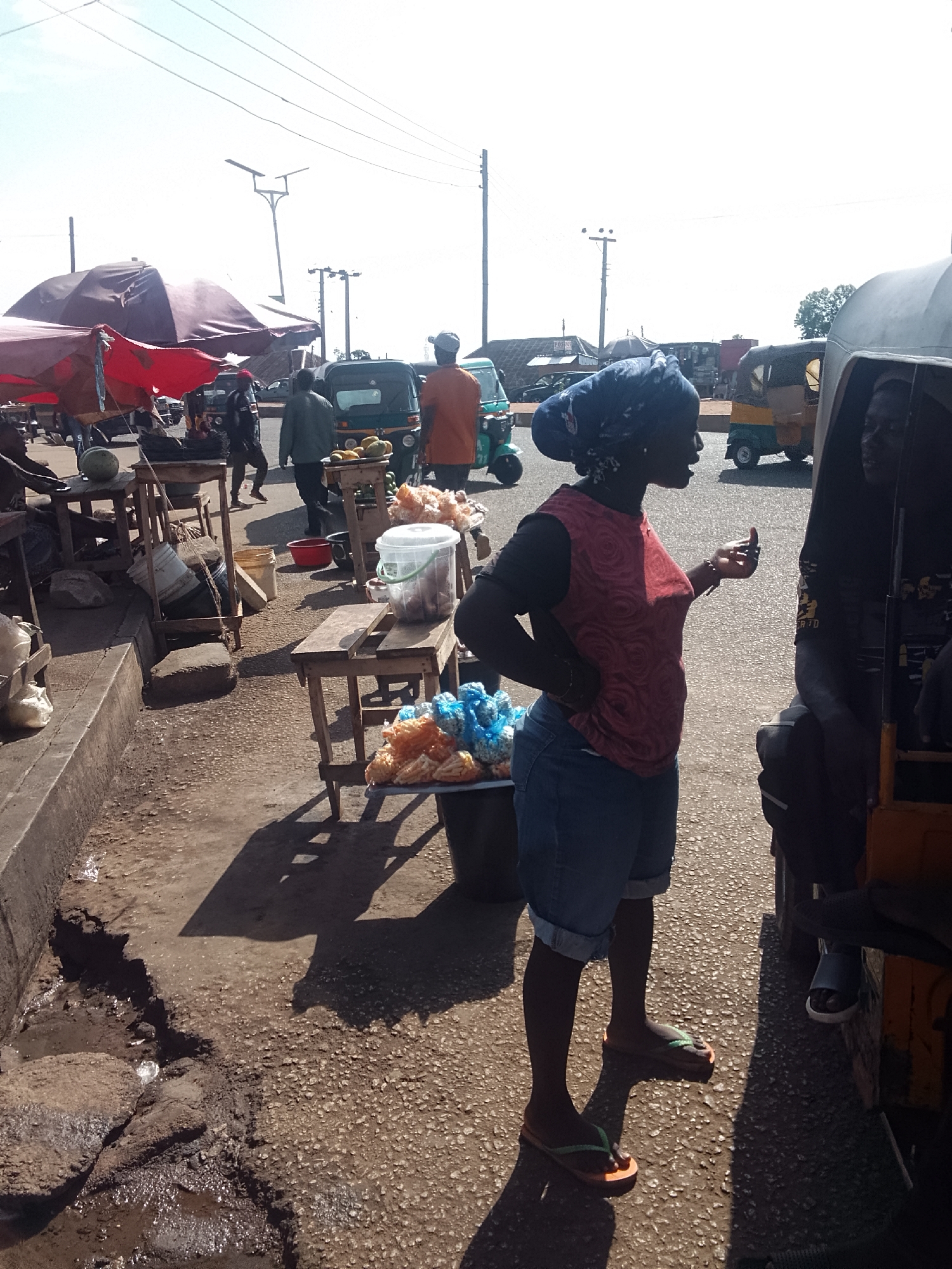 The image shows an outdoor street scene in a city location of Unknown, Unknown. The scene includes a woman wearing a red shirt, denim shorts, and a headwrap, standing near a motorized vehicle (likely a tuk-tuk or auto-rickshaw). The woman appears to be in conversation with someone inside the vehicle.
Behind the woman, there are makeshift stalls or tables displaying goods, including what appears to be food items in plastic bags. Other individuals can be seen walking or standing in the background. The location seems to be a busy street with other tuk-tuks and various commercial activities in the vicinity. Power lines are visible overhead, and the weather appears to be sunny. The condition of the road and pavement suggests it is a somewhat urban or peri-urban environment.