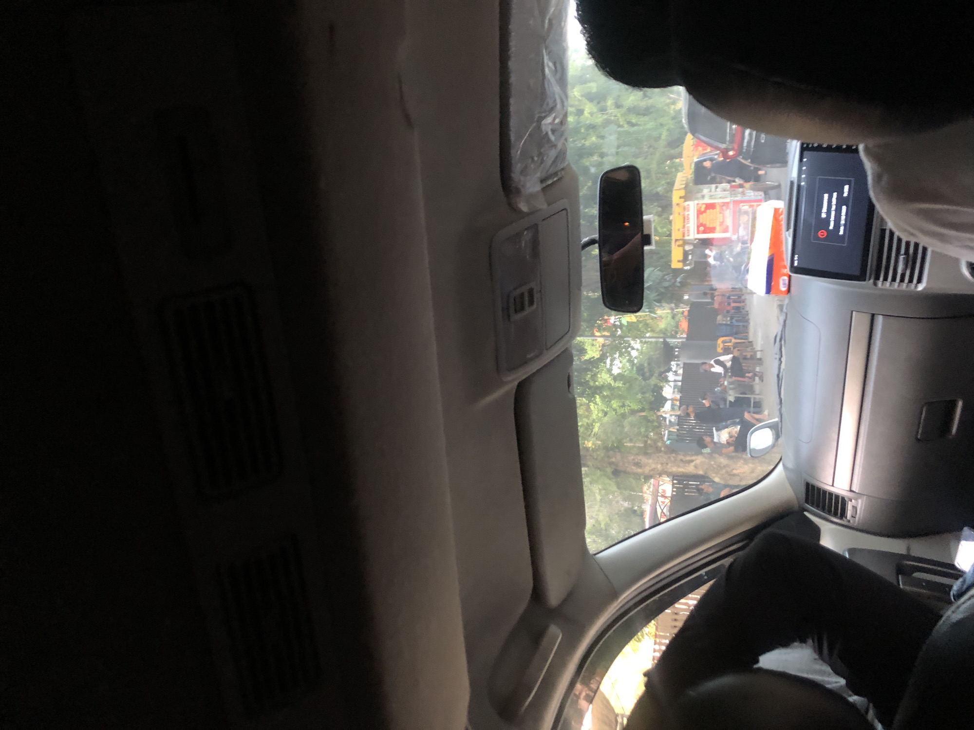 The image, taken from inside a vehicle in what appears to be the city of Medan, Indonesia, shows a view through the windshield. The foreground is dominated by the interior of the car, including the dashboard, a rearview mirror, and the heads of passengers. Outside the vehicle, a street scene unfolds with people gathered around food stalls. Trees and a fence line the background, suggesting a casual outdoor setting within the urban environment. The overall impression is one of a typical day in a bustling Indonesian city, possibly during daytime hours given the natural light.