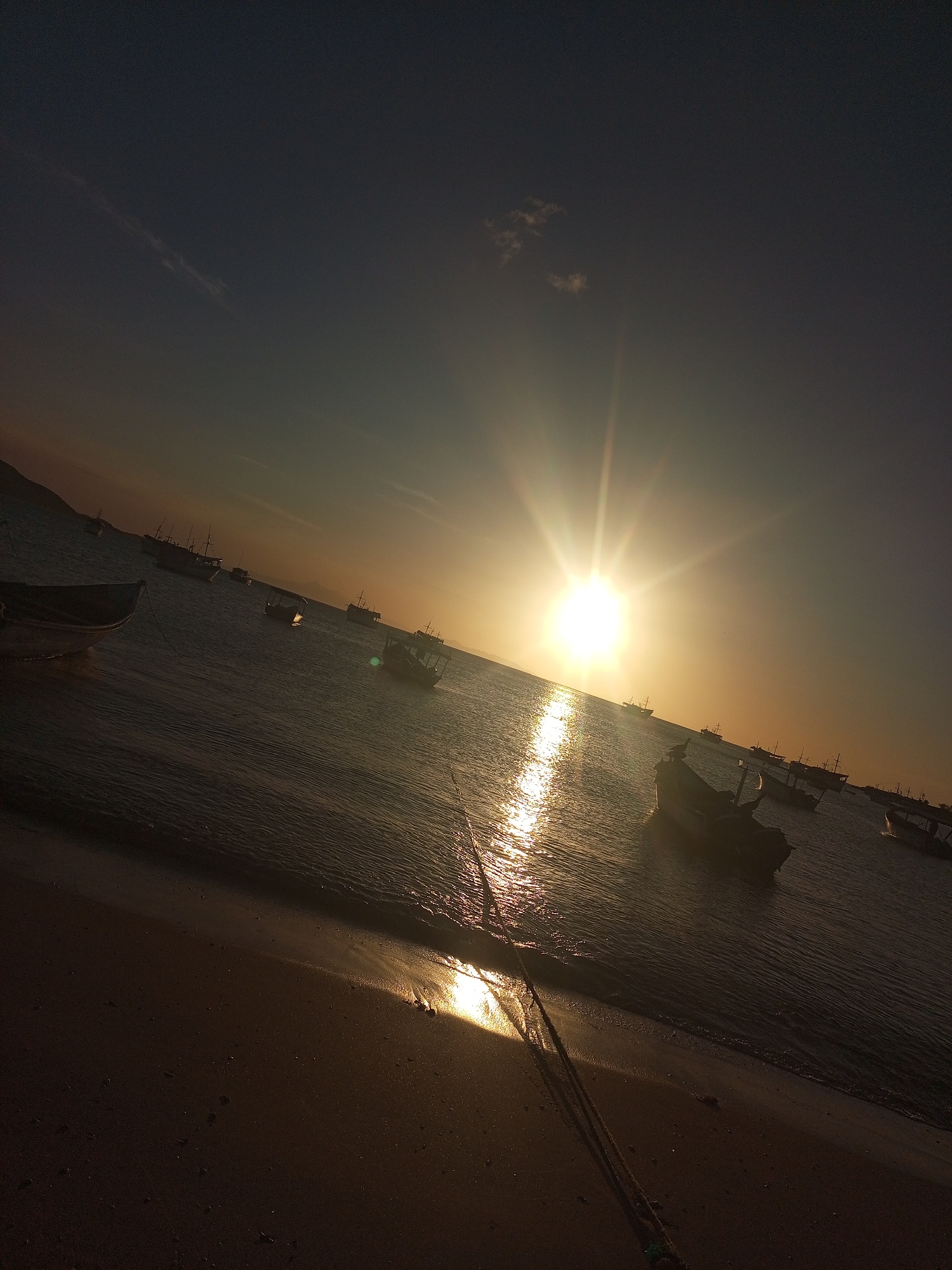 The image depicts a coastal scene in Juan Griego, Venezuela, during either sunrise or sunset. The sun, a bright luminous sphere with a prominent starburst effect, is positioned in the mid-right portion of the frame, casting a strong golden-orange glare across the calm water. The sky transitions from a dark blue-grey in the upper regions to a warmer, brighter hue near the horizon. Faint, wispy clouds are visible in the darker sky overhead.
Approximately ten to fifteen boats of varying sizes are visible on the water, appearing as dark silhouettes against the sun's reflection. A prominent, darker boat is positioned in the lower-left quadrant, partially resting near or on the sandy shore. A dark rope or line extends diagonally from the lower-left corner of the frame, across the wet, dark brown sand of the immediate foreground, and into the water, with its connection point obscured. Gentle wavelets are observed along the narrow strip of sandy shoreline. A dark, undulating landmass is silhouetted against the horizon on the far left. No individuals are present in the scene.
