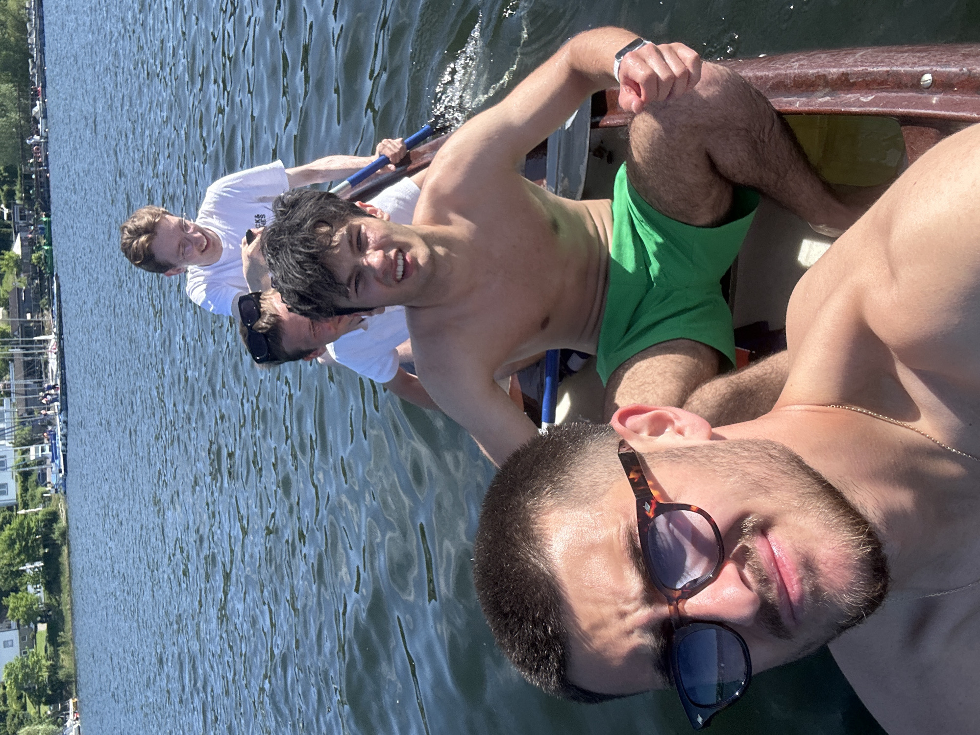 Four men are in a rowboat on the Möhnesee, Germany. The photo is taken from the front of the boat, with a man wearing sunglasses in the foreground. Behind him, a man with green shorts and no shirt is sitting. Further back, two men are rowing, wearing white t-shirts. The water of the lake surrounds the boat, with trees and buildings visible on the shoreline.