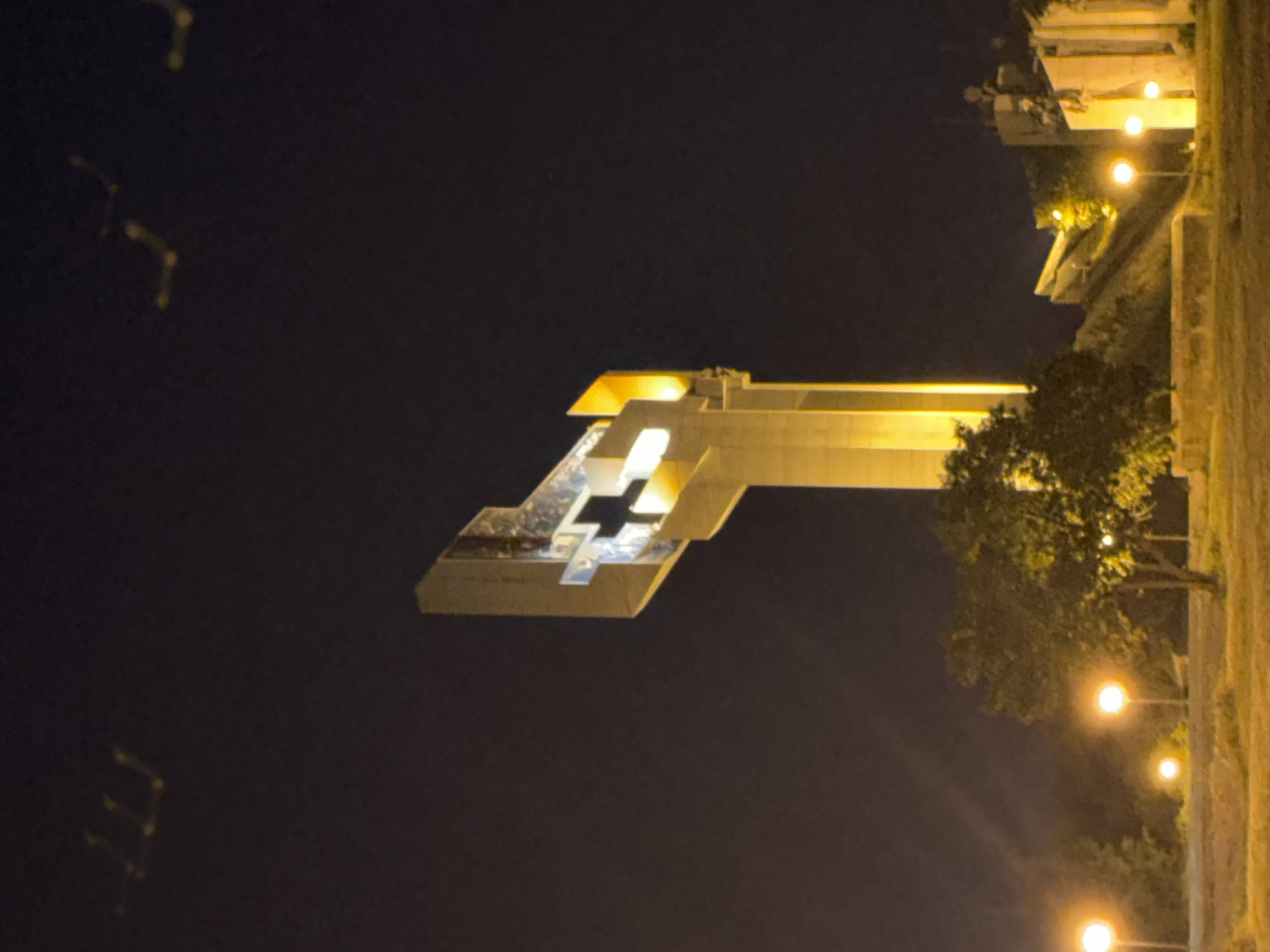 At night, the Defenders of Stara Zagora Memorial Complex, Bulgaria is illuminated and visible. The memorial is composed of layered horizontal slabs, with a cross-shaped void. The monument is situated next to a line of street lamps illuminating a path lined with trees and a stone wall. There are a few other light sources on the left side of the image, but they appear to be distant.
