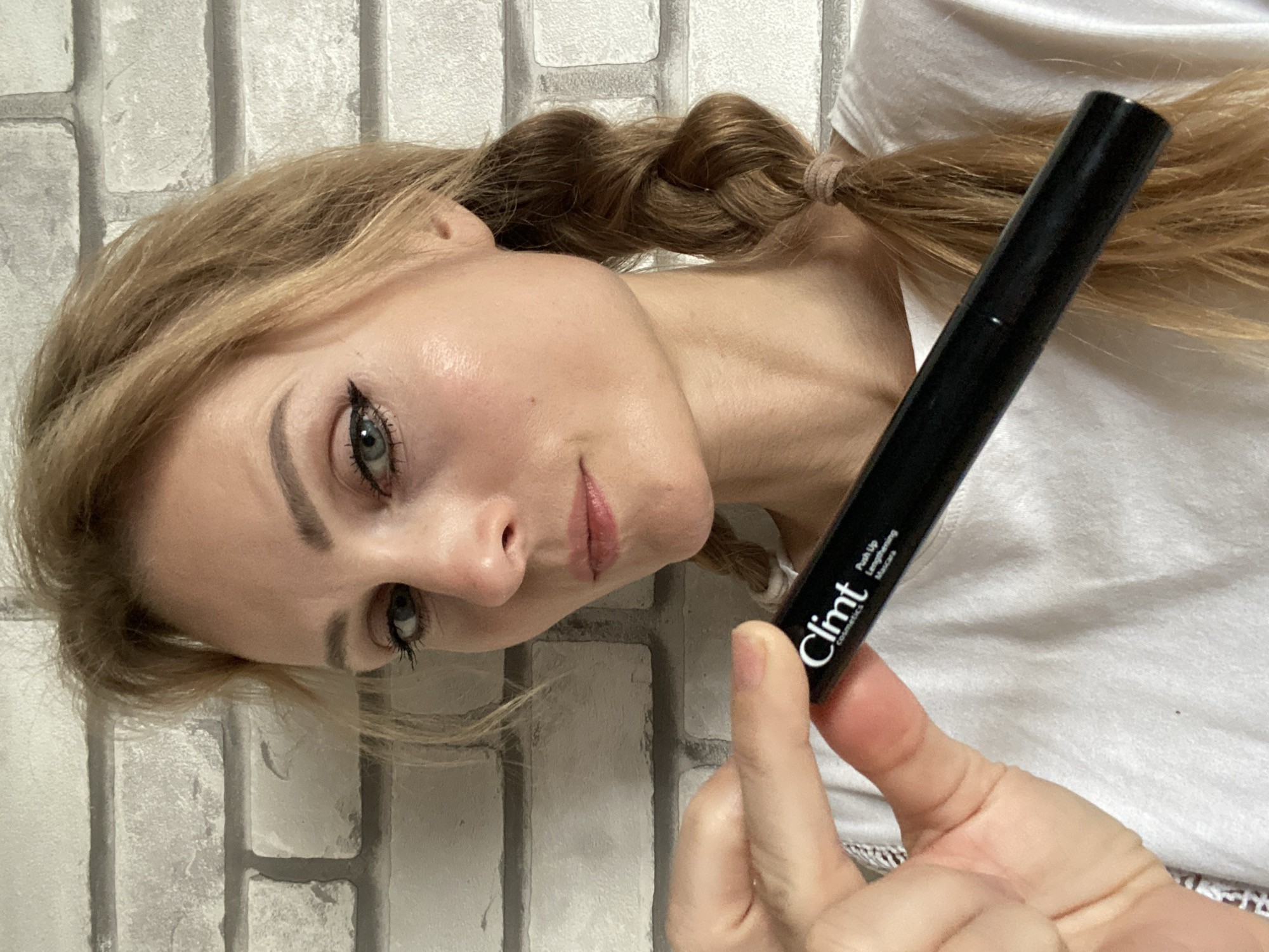 A fair-skinned woman with blonde hair braided into pigtails stands against a grey brick wall. She holds a black mascara tube with a white label that reads "Climt cosmetics Push Up Lengthening Mascara" in her left hand, in front of her chest. She is wearing a white t-shirt. Her makeup includes winged eyeliner and lip gloss. The image appears to be a selfie. The location is not specified in the image, therefore the city of Міханавіцкі сельскі Савет, Belarus cannot be determined.