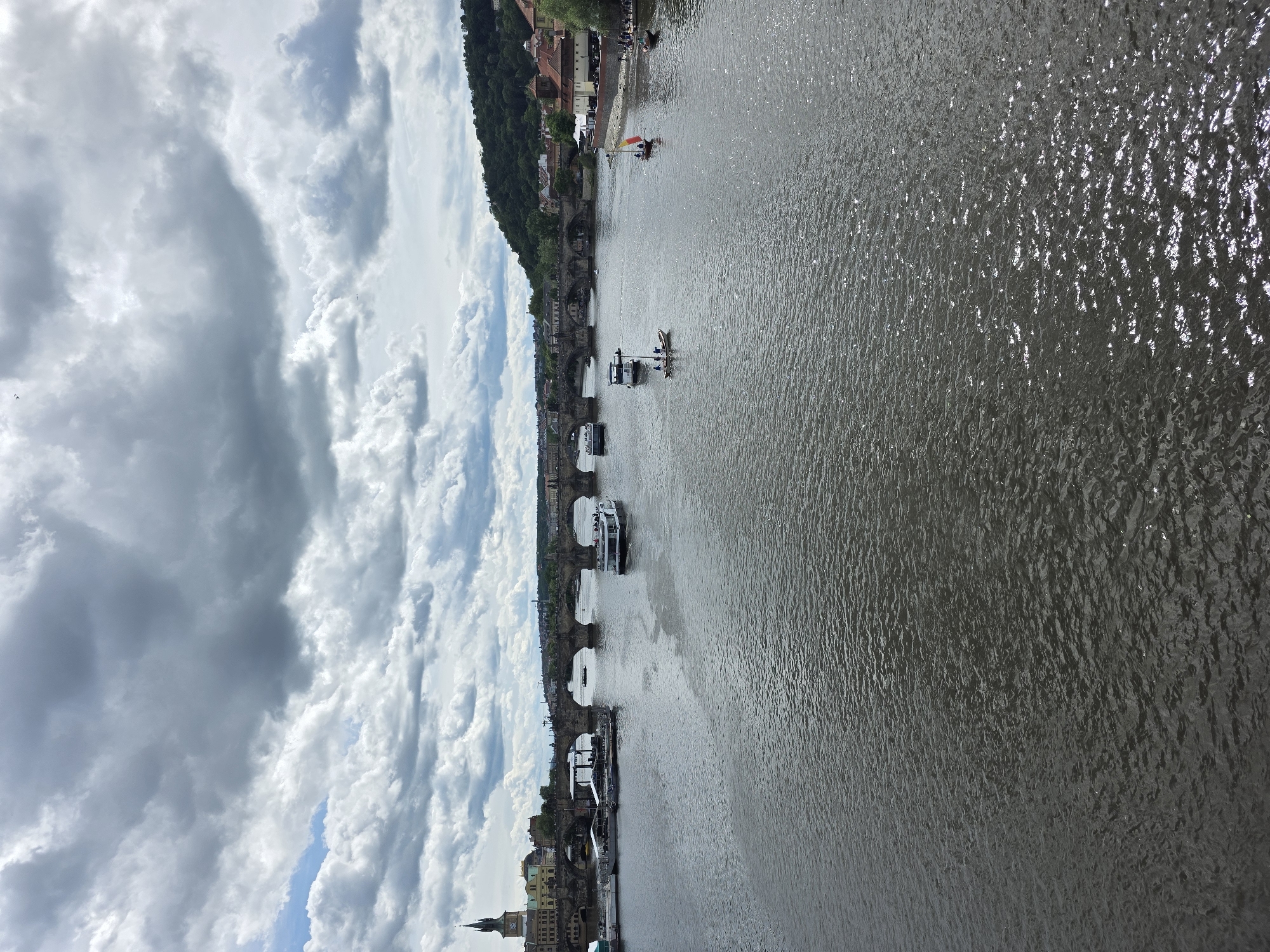 The image captures a wide outdoor scene featuring a river, a historic bridge, a city along the riverbank, and a cloudy sky. The perspective is from an elevated viewpoint, looking down and across the river.
The river, occupying the right half of the frame, shows textured ripples on its surface, reflecting daylight. Several boats are visible on the water. Closest to the foreground, a large, modern white passenger boat moves away from the viewer, situated near the bridge. Further upriver, a smaller, dark-hulled vessel is also seen, and even further, a small boat with a distinct red and yellow flag or sail, possibly carrying passengers. A small kayak or canoe with two people onboard is positioned near the larger white boat.
A multi-arched stone bridge spans the river from left to right, connecting the visible city bank with what appears to be another bank or the continuation of the city. Structures, possibly bridge towers or statues, are discernible on the bridge.
On the left side of the river, a vibrant cityscape unfolds. A steep, tree-covered hillside rises behind the buildings, with some red-roofed structures nestled into the greenery. Along the immediate riverbank, a cluster of traditional buildings with varying architectural styles and roof colors lines the water's edge. In the lower left corner, a prominent clock tower with a pointed spire stands tall among other buildings.
The sky, filling the upper portion of the frame, is largely overcast with large, fluffy white and grey clouds, though patches of blue sky are visible between them. The overall lighting suggests it is daytime. No discernible text is visible in the image. The scene conveys a sense of historical charm and natural beauty, possibly a European city.