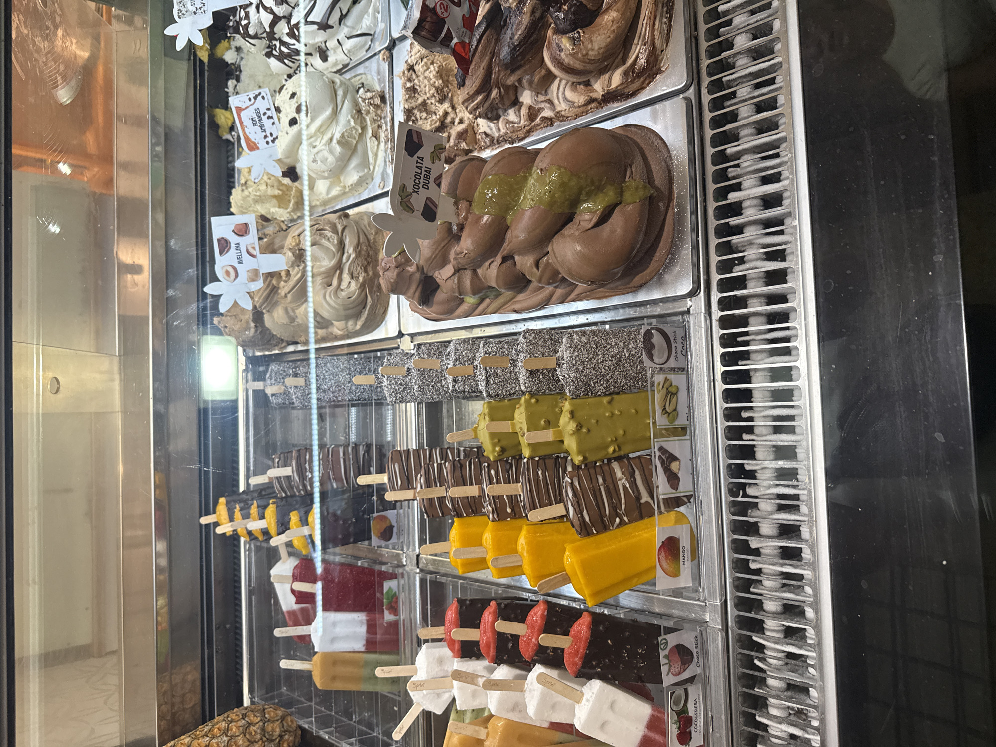 This image depicts an ice cream display case at a shop in Tossa de Mar, Spain. The case is filled with a variety of colorful gelato and popsicle flavors. In the foreground, several rows of popsicles are visible, including some with dark chocolate coating and what appear to be pistachio nuts, as well as some with yellow and red fruit flavors. Behind the popsicles, tubs of gelato are displayed, with flavors like chocolate, vanilla, and a pistachio-swirled variety. Small signs with flavor names in Italian are placed in some of the gelato tubs, such as "AVELLANA" (hazelnut) and "XOCOLATA DUBAI". The overall environment appears to be a commercial establishment with a refrigerated display case.