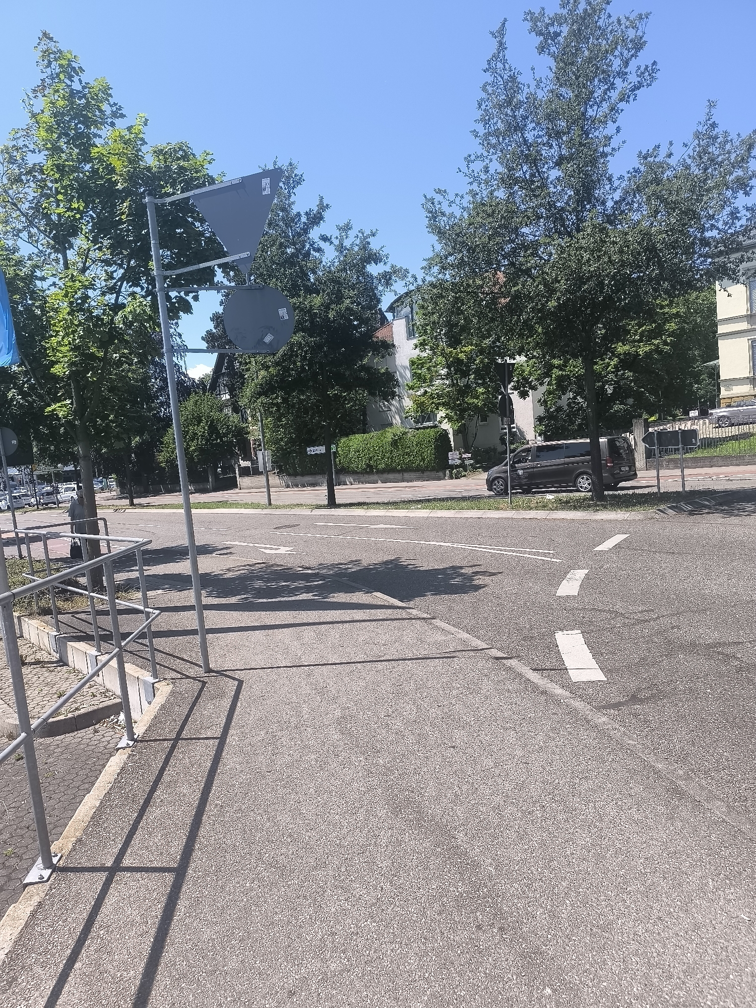 The image shows a street scene on a sunny day. In the foreground, a grey asphalt road is visible with white road markings indicating a crosswalk. To the left, a metal railing and a concrete curb line the sidewalk, which is paved with small grey stones. Beyond the curb, a few parked cars are visible. In the middle ground, a dark-colored minivan is driving on the road. A metal pole with two traffic signs, one triangular and one circular, stands near the road. Lush green trees with dense foliage line the street, casting shadows on the asphalt. In the background, buildings with light-colored facades and pitched roofs are visible, interspersed with more greenery. The sky is clear and bright blue.