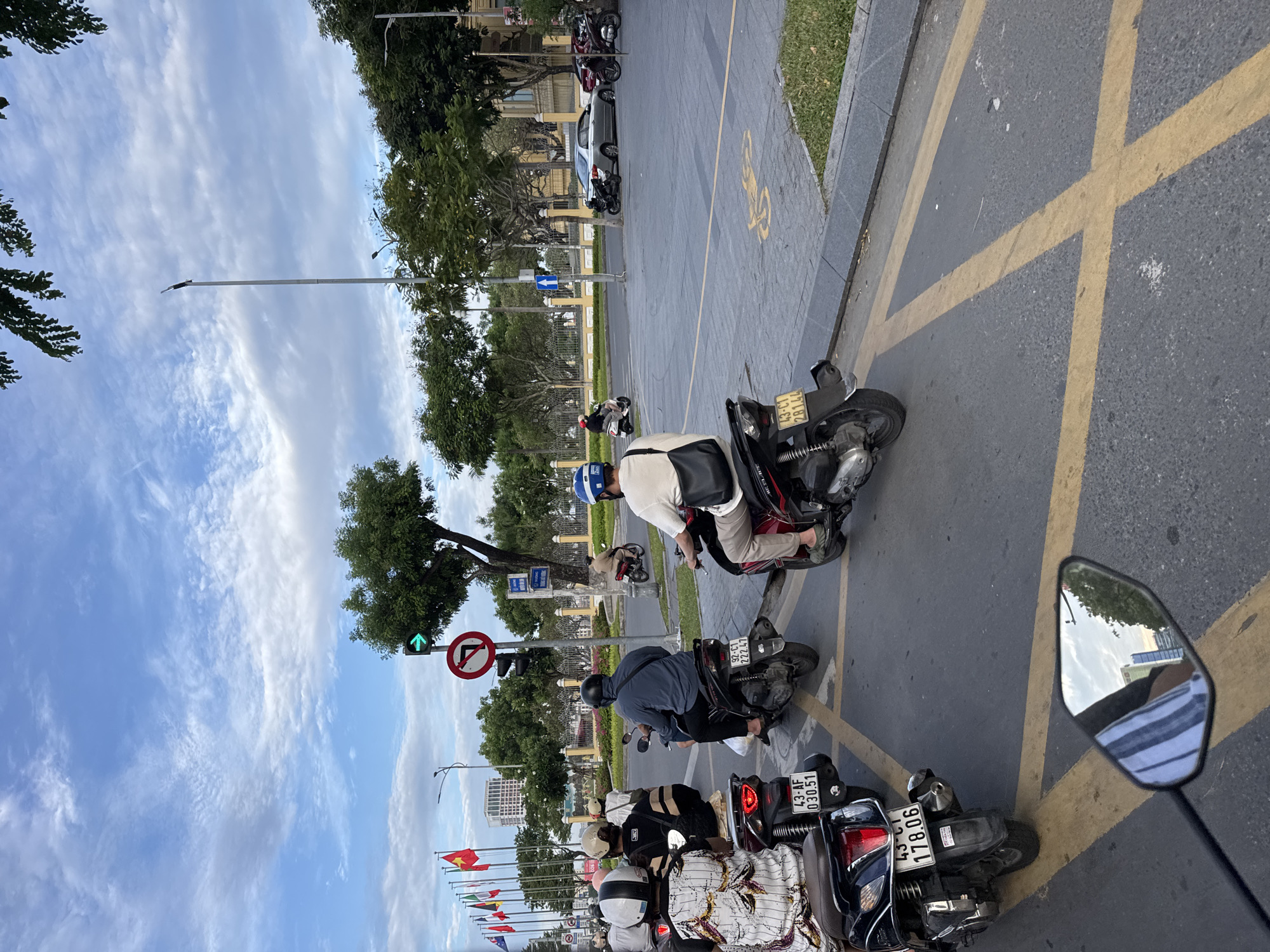 The image captures a street scene in Da Nang, Vietnam, likely near a major intersection given the presence of traffic signals and road markings. Several motorcycles, a common mode of transport in Vietnam, are visible, carrying individuals wearing helmets and casual attire. One motorcycle in the foreground has the license plate "43-AF 030.51" and another reads "43-CT 281.44". Another motorcycle in the left foreground has a license plate "43-CI 178.06". In the background, traffic lights show a green arrow indicating a turn, and a red circular sign with a diagonal line indicates a prohibition. Flags are seen on poles along the roadside, suggesting a potentially official or significant area. The environment appears urban, with buildings and trees lining the street, under a sky with scattered clouds. The road is paved and marked with yellow lines, indicating traffic lanes and potentially a no-parking zone with a bicycle symbol painted on the ground. The overall atmosphere suggests moderate traffic flow in an outdoor urban setting.