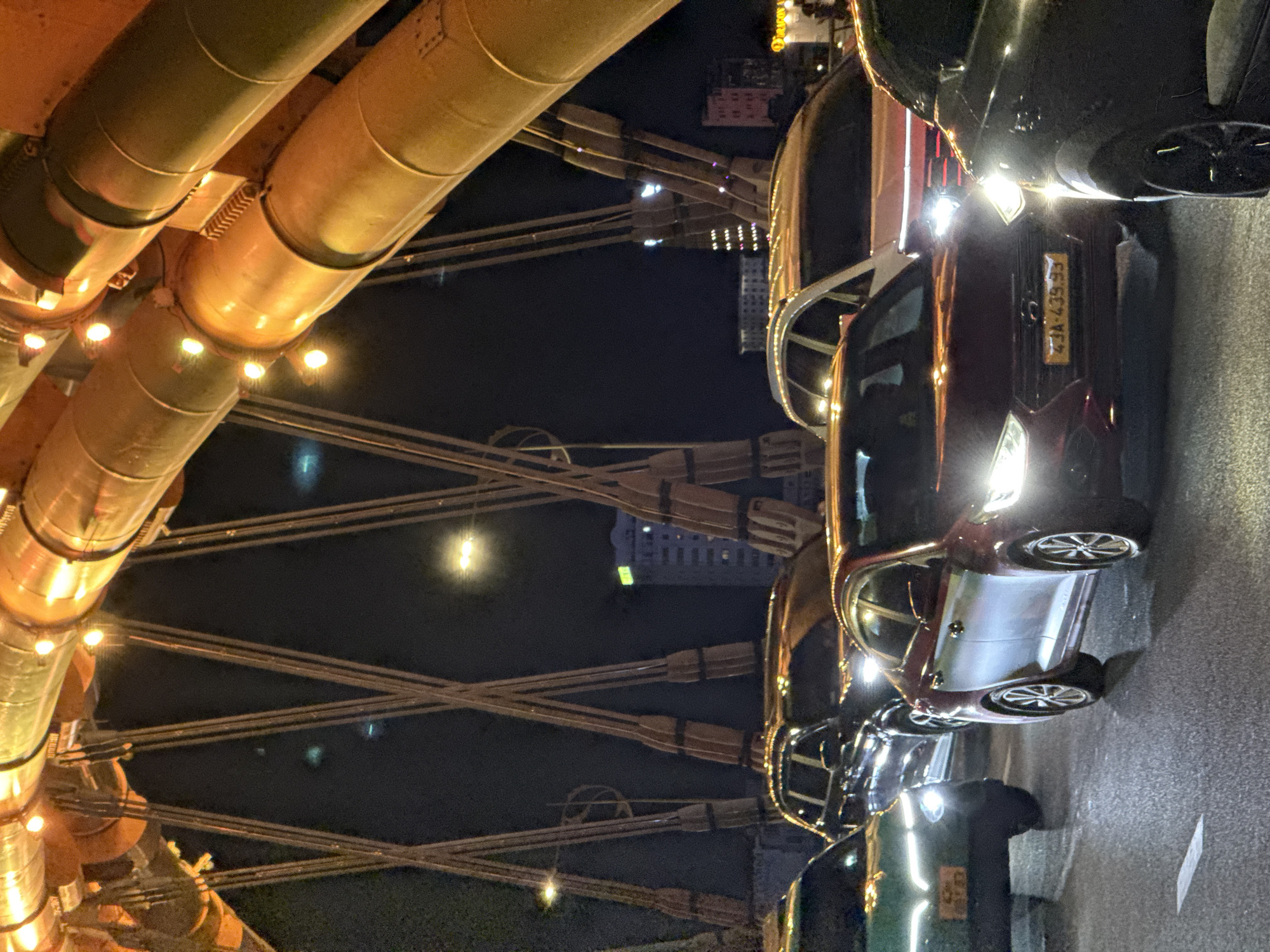 This nighttime photograph depicts a traffic scene on the Dragon Bridge in Đà Nẵng, Vietnam. The upper and right portions of the frame are dominated by the bridge's large, tubular, golden-orange arched structural elements, which are illuminated by a series of embedded circular lights. Dark, multi-stranded suspension cables extend downwards from these arches, creating diagonal patterns against the dark sky.
On the bridge deck below, numerous vehicles are observed in traffic. A dark red Hyundai sedan, displaying the Vietnamese license plate "43A-439.53," is prominently featured in the mid-ground, facing slightly left with its LED headlights illuminated. To its immediate left, a dark green VinFast sedan is partially visible in the foreground, also with its headlights on. Behind these, a dark SUV is discernible, and further to the right, a light-colored SUV is partially visible. Other vehicles with illuminated headlights and taillights are present in the background, indicating congested traffic conditions. The asphalt road surface is visible in the foreground, featuring a faint white lane marking. In the distant background, dimly lit urban buildings stand against the dark night sky, marked by scattered artificial lights. No individuals are distinctly visible.