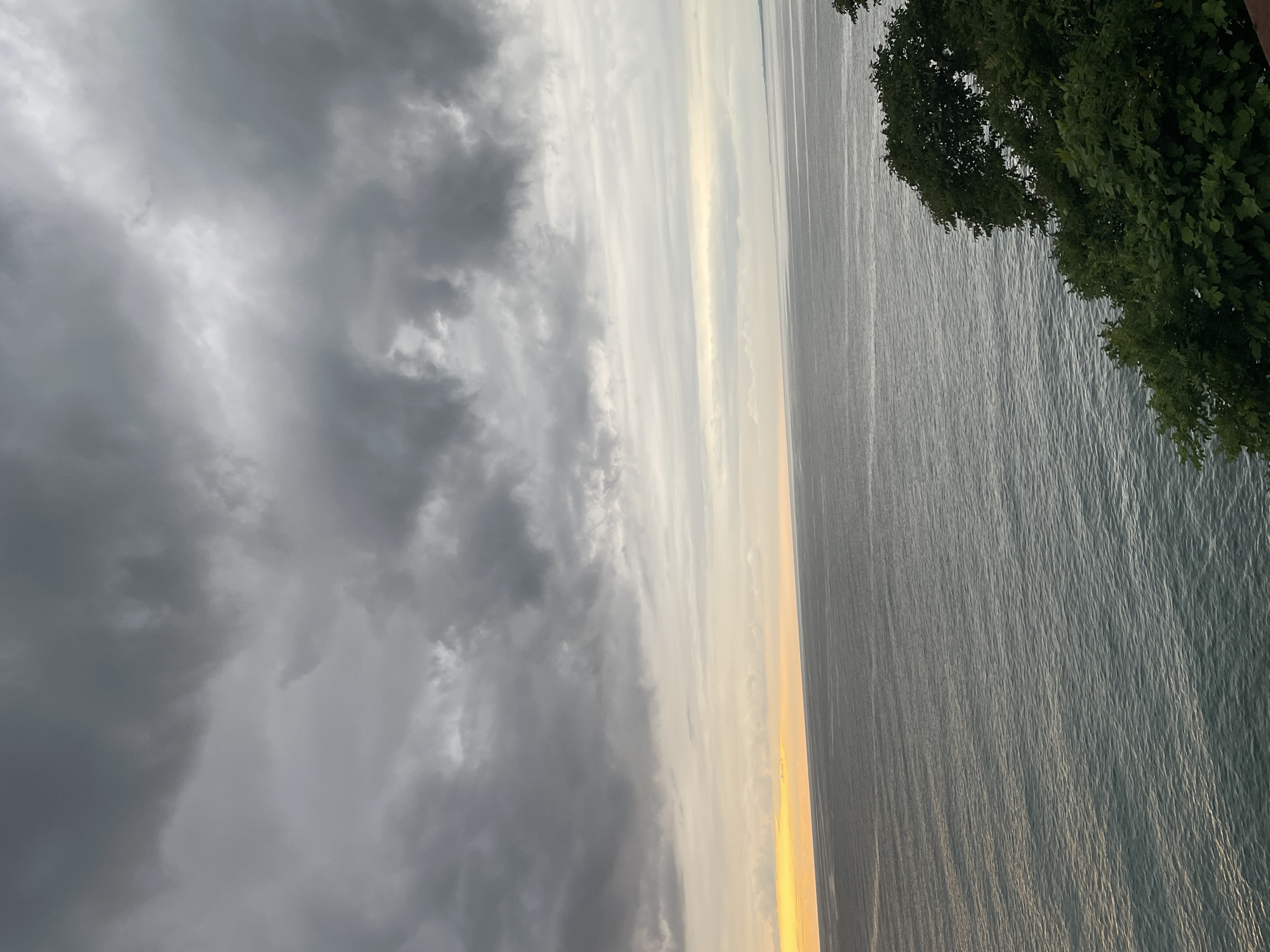 The image shows a body of water, likely the Black Sea, with a dramatic cloudy sky. The clouds are dark and heavy, with some lighter areas suggesting the sun is trying to break through. The water is calm with small ripples. In the foreground, on the right side of the image, there is a lush green tree line, indicating a coastal environment. The overall mood is overcast and potentially stormy, but with a hint of light on the horizon. The scene is captured from an elevated perspective, overlooking the water. There are no people or man-made structures visible in the image, suggesting a natural, possibly remote, coastal area. The location is specified as Buknari, Georgia, which is a coastal settlement known for its beaches.