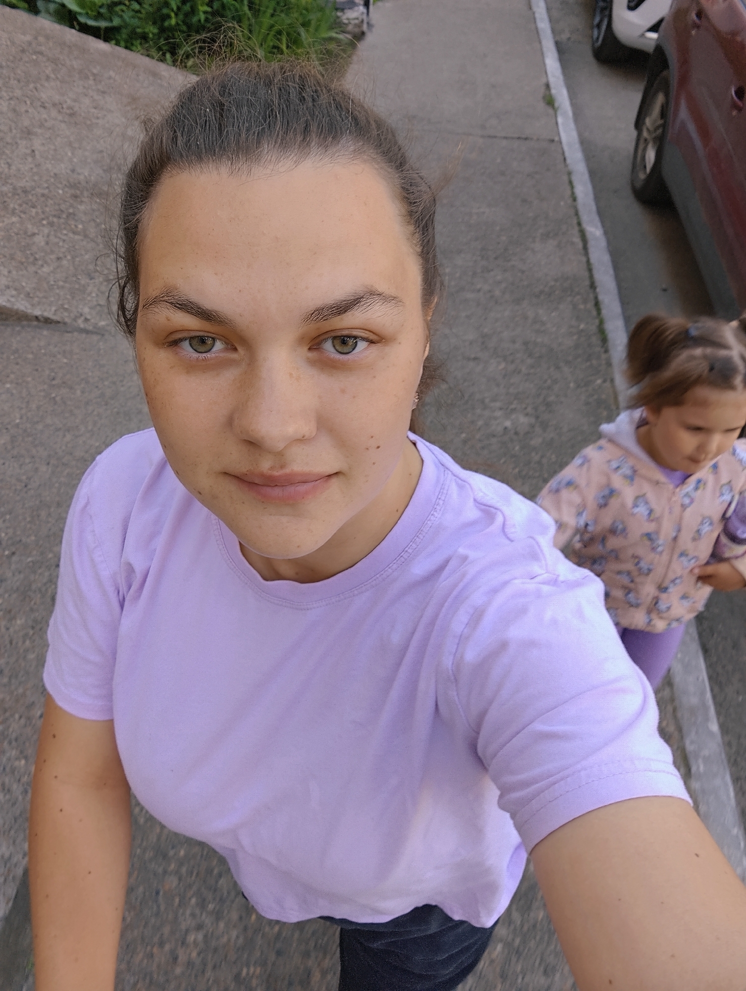 A woman and a young girl are standing on a sidewalk in Chaikovsky, Russia. The woman is in the foreground, looking at the camera, and is wearing a light purple t-shirt. The girl is behind her, to the right, and is wearing a light pink jacket with a unicorn pattern and light purple pants. Two cars are parked on the street to the right of the sidewalk. The sidewalk is made of concrete.