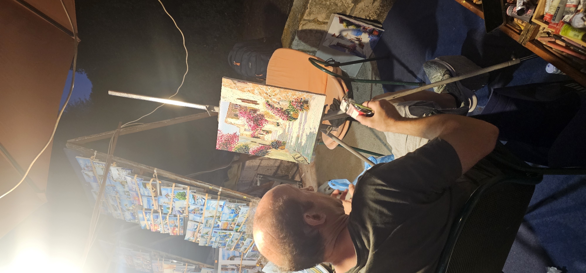 The image depicts an artist painting in Grad Supetar, Croatia, at nighttime. The scene is lit by a bright artificial light source, likely a lamp, positioned above an easel. The artist, a middle-aged man with a bald head, is seated and focused on a painting of a picturesque European street scene, characterized by buildings adorned with vibrant pink flowers.
The easel is set up outdoors, under a large umbrella or awning, suggesting a market or exhibition stall. To the left of the easel, a rack displays numerous smaller paintings, possibly postcards or prints, depicting similar scenic views. The background is dark, hinting at the late hour, with hints of foliage and stone structures.
The artist is holding a brush and appears to be actively engaged in his work. He is dressed in casual attire. The overall atmosphere is one of focused creativity and artistic display. The presence of the artwork and the artist's attire suggest an outdoor art sale or demonstration. The lighting casts distinct shadows, further emphasizing the nighttime setting. No visible text is present in the image.